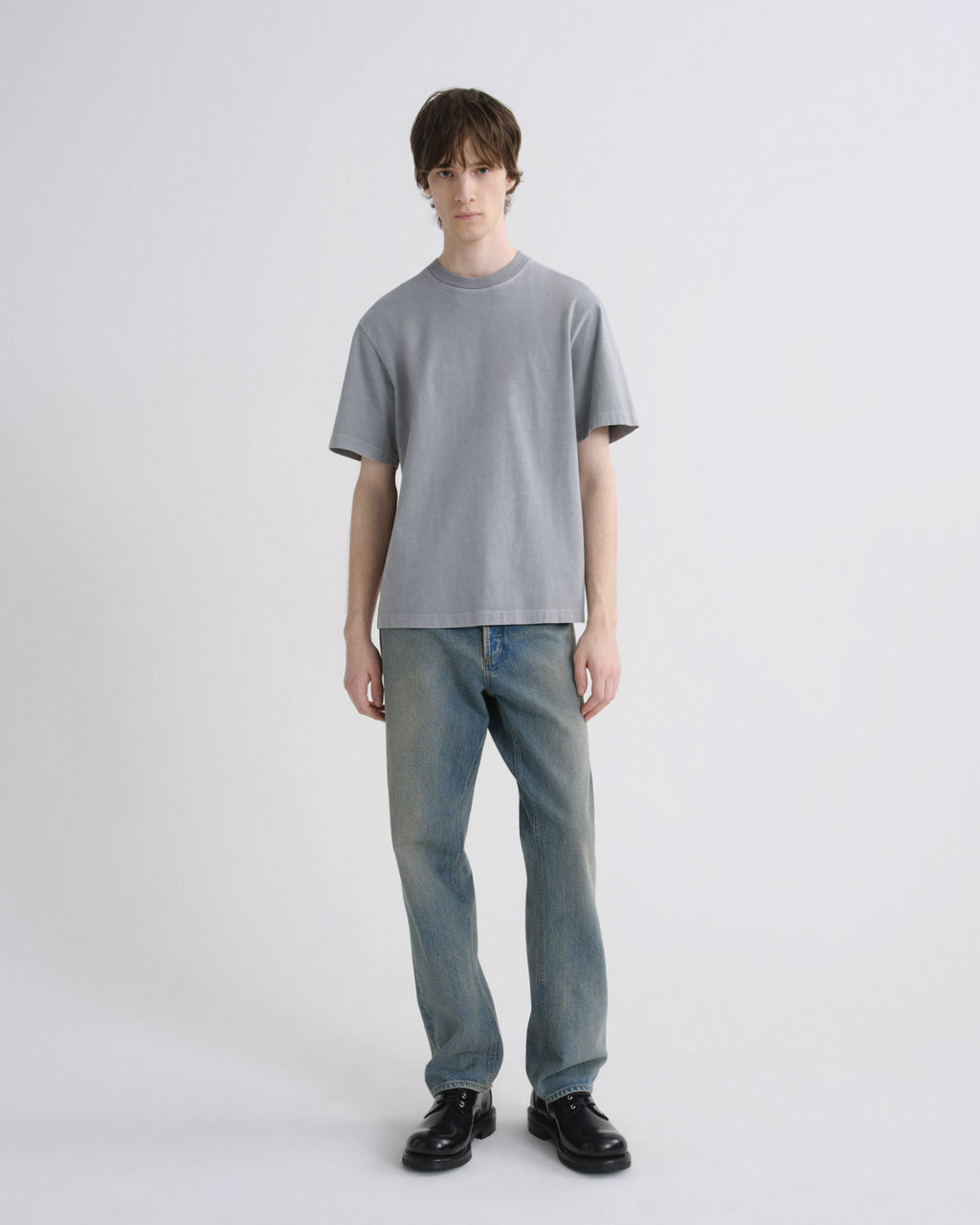 A model stands against a light gray studio background wearing a light gray crewneck t-shirt, light blue jeans, and black leather shoes.