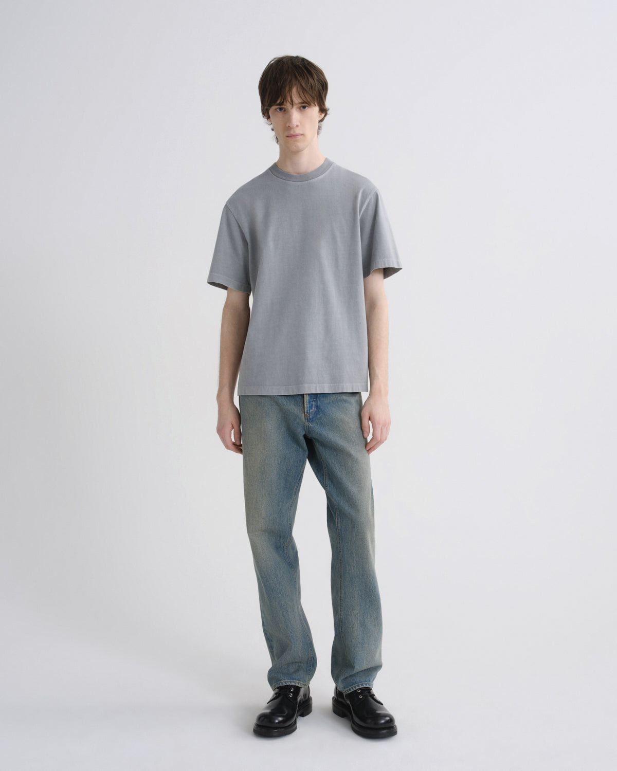 A model stands against a light gray studio background wearing a light gray crewneck t-shirt, light blue jeans, and black leather shoes.