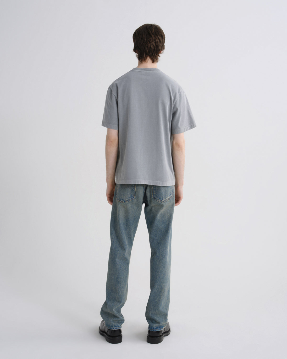 A man viewed from behind wearing a light gray short-sleeve t-shirt and light blue jeans, standing against a plain light gray studio background.
