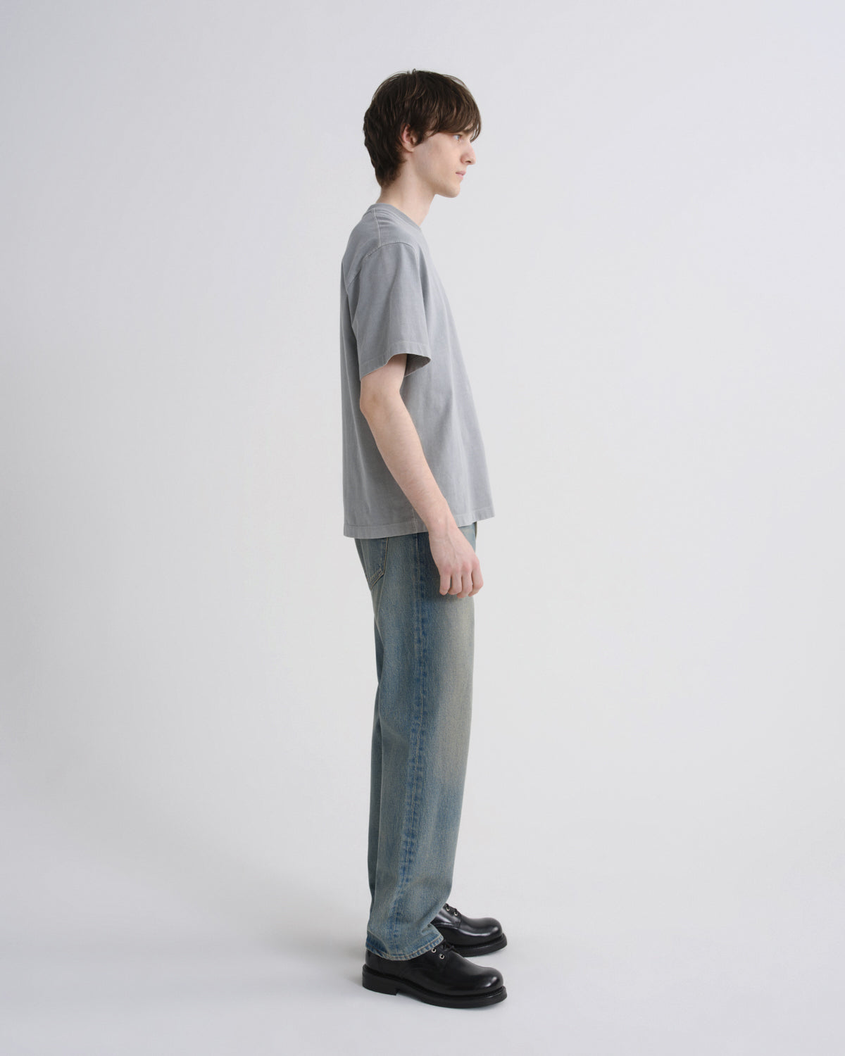 A model standing in profile wearing a light gray oversized short-sleeve t-shirt, light-wash blue jeans, and black leather shoes against a plain light gray studio background.