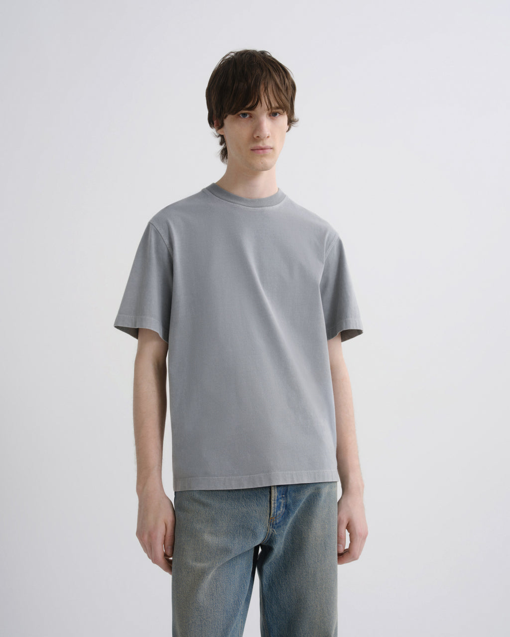 A young man modeling a light gray crew-neck t-shirt and blue jeans against a plain light gray studio background.