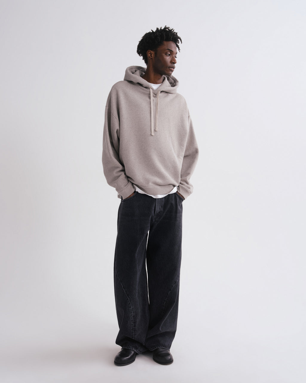 A man wearing a light beige oversized pullover hoodie with a gray inner hood lining, dark wide-leg jeans, and black shoes, standing against a plain light gray studio background.