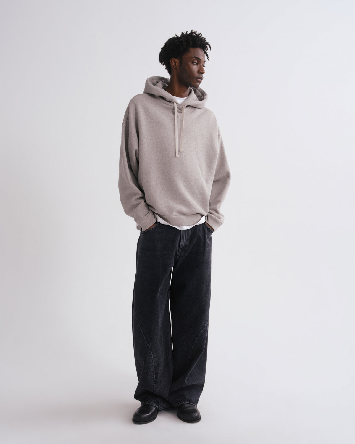 A man wearing a light beige oversized pullover hoodie with a gray inner hood lining, dark wide-leg jeans, and black shoes, standing against a plain light gray studio background.