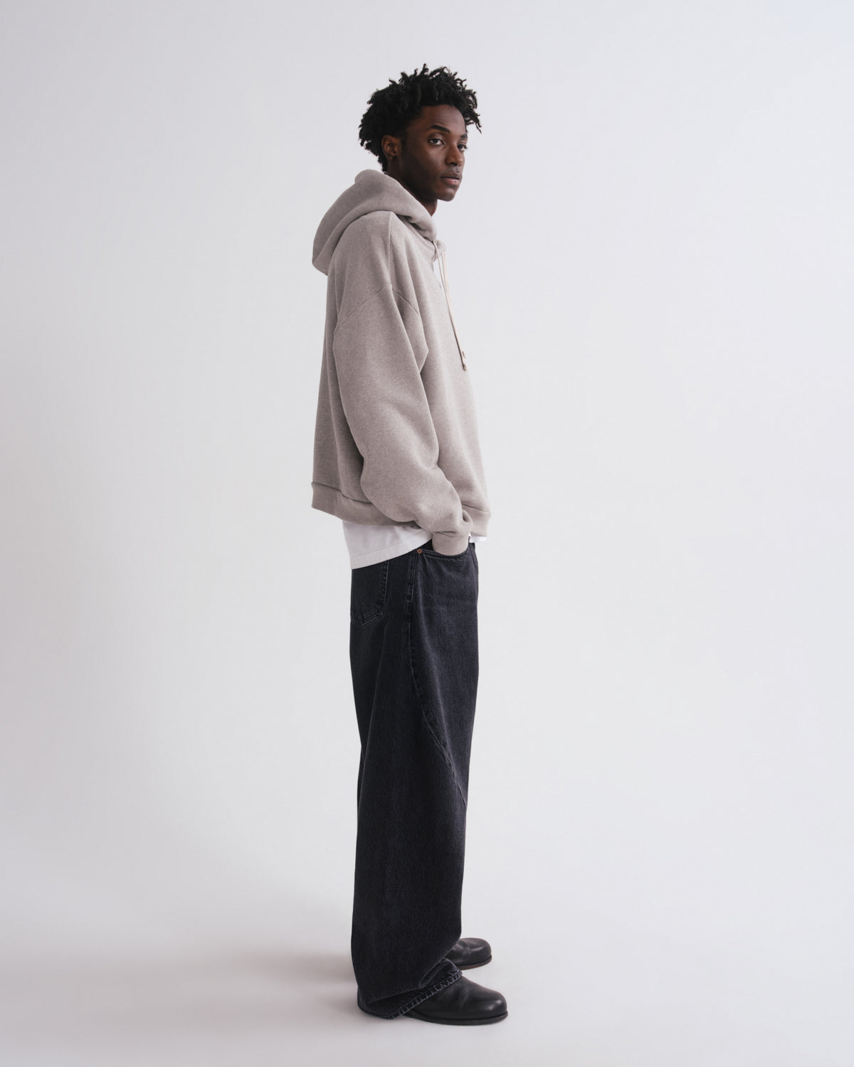 A man stands in profile wearing a light beige oversized hoodie, dark wide-leg jeans, and black shoes against a plain light gray studio background.