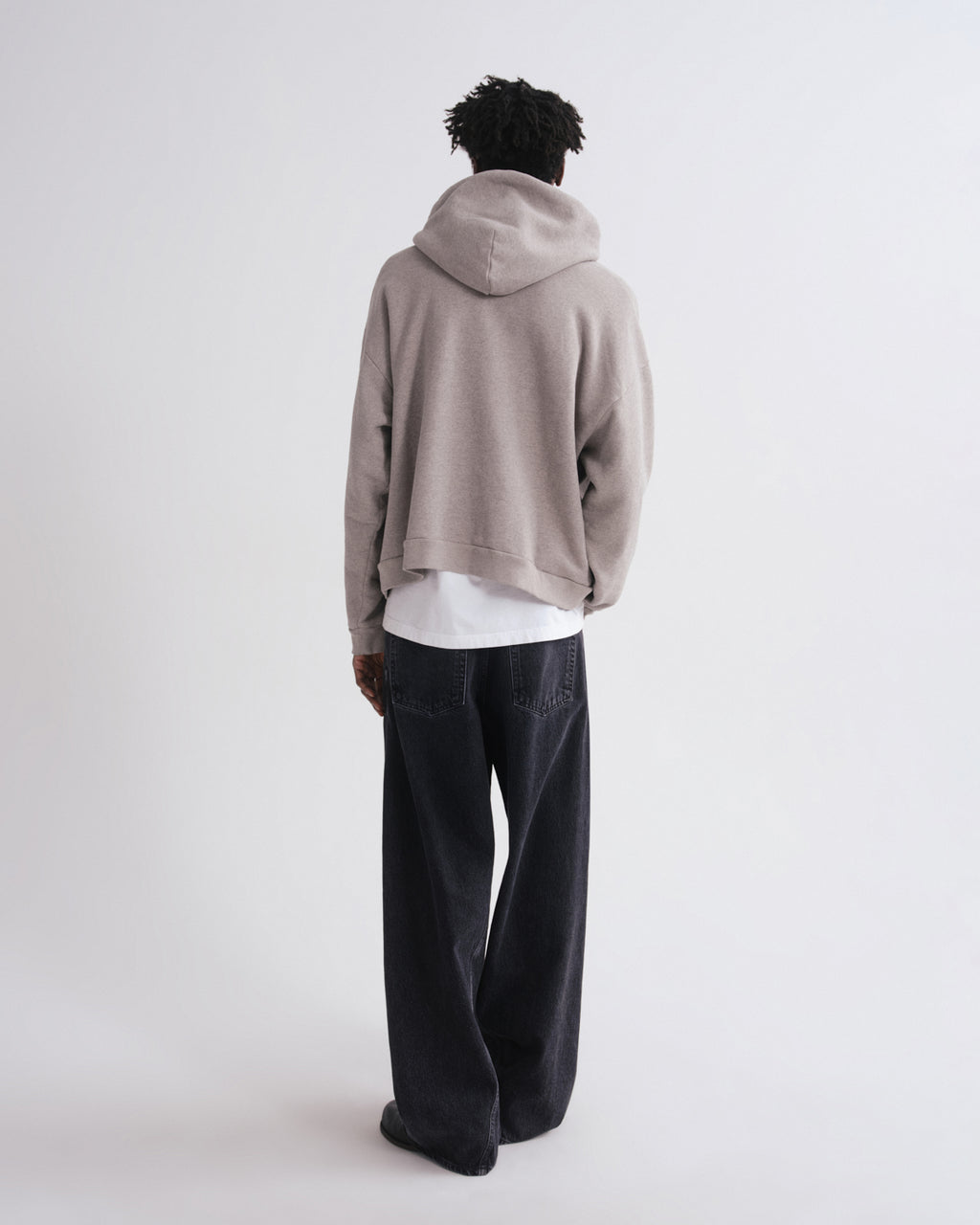 A person shown from the back wearing a cropped taupe-beige pullover hoodie over a white shirt and dark charcoal wide-leg jeans, photographed against a seamless light gray studio background.