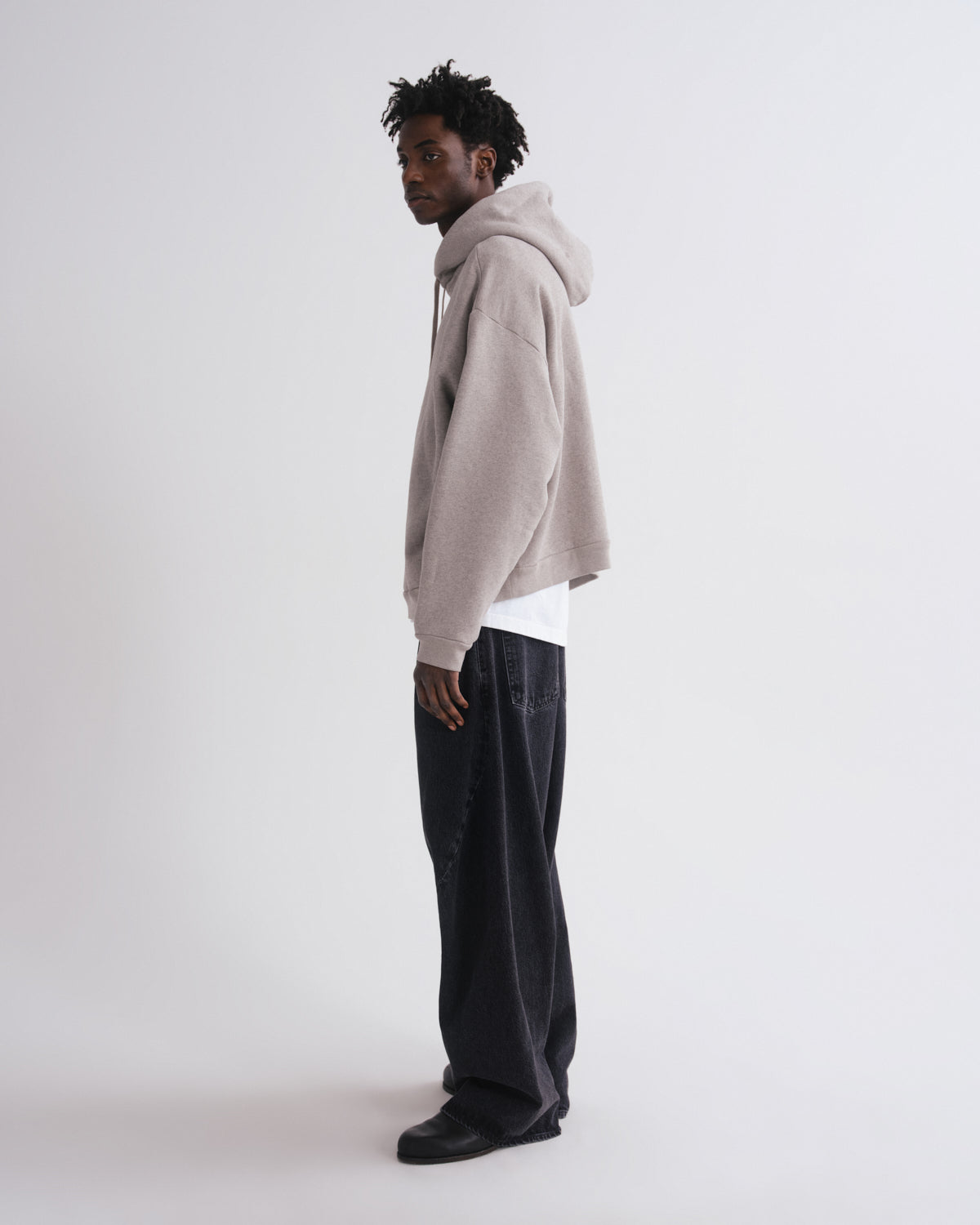 A man stands in profile wearing a light beige oversized cropped hoodie, a white undershirt, dark charcoal wide-leg jeans, and black shoes against a plain light gray studio background.