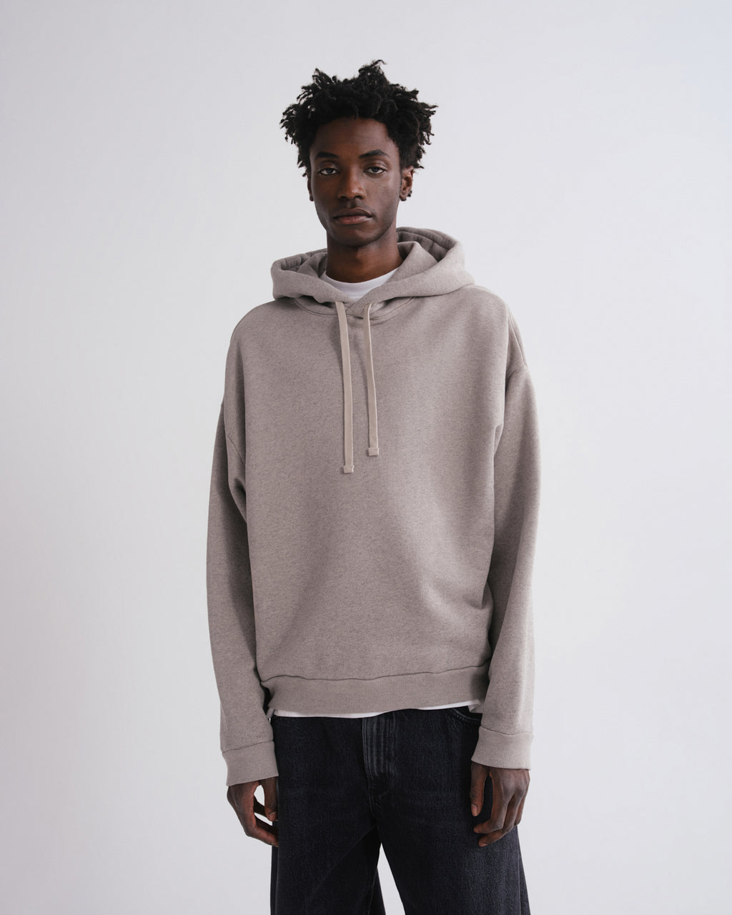 A man wearing a light taupe-beige pullover hoodie with matching drawstrings, layered over a white undershirt and dark jeans, standing against a plain light gray studio background.