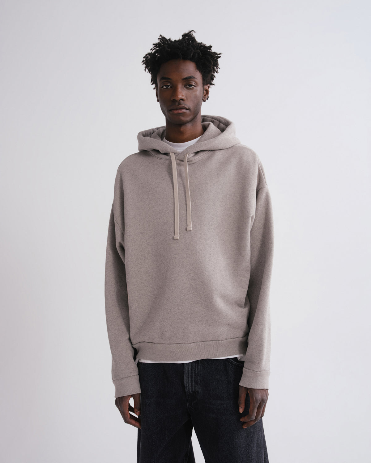 A man wearing a light taupe-beige pullover hoodie with matching drawstrings, layered over a white undershirt and dark jeans, standing against a plain light gray studio background.