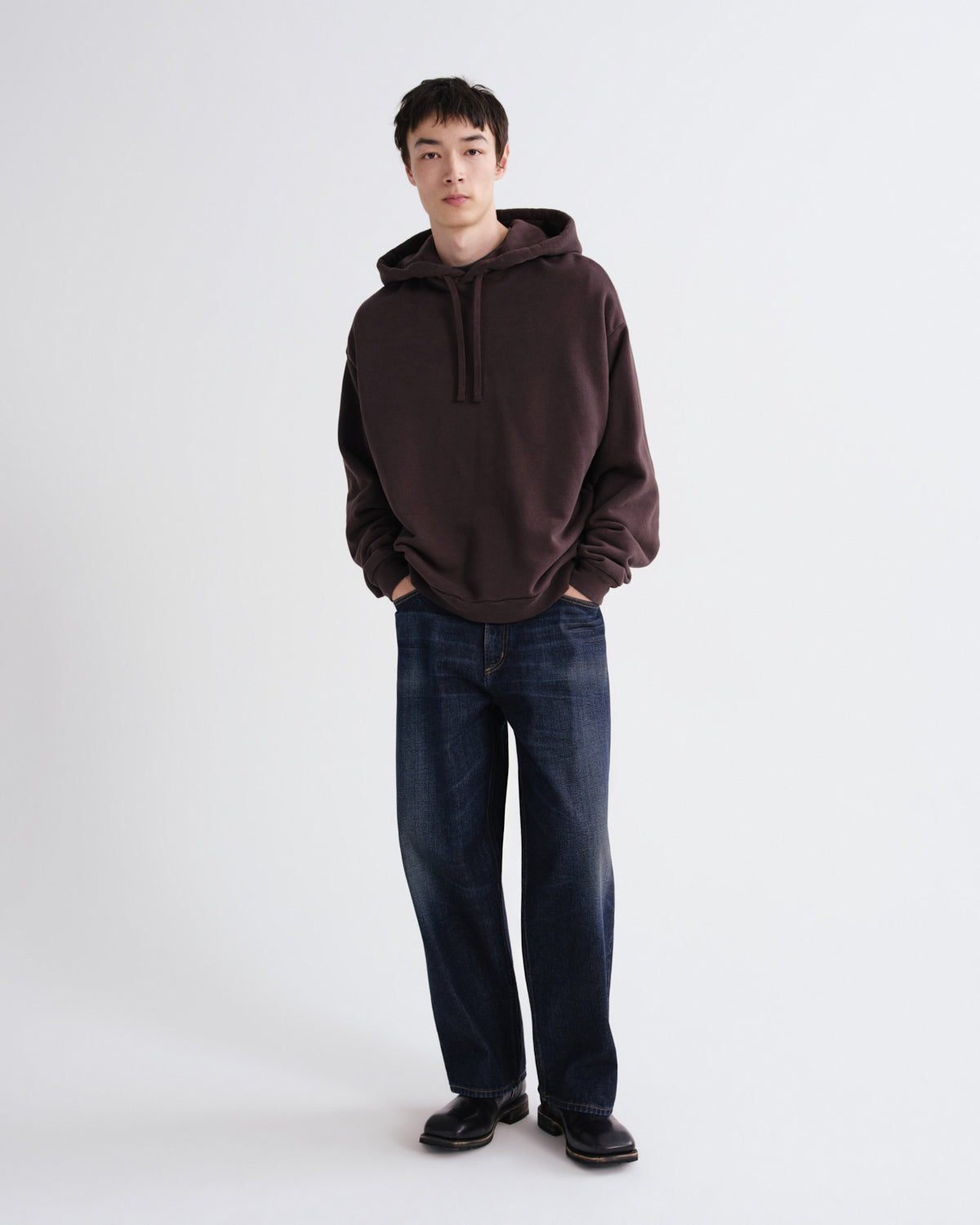 A man wearing a dark brown pullover hoodie, dark blue jeans, and black shoes, standing against a plain light gray studio background.