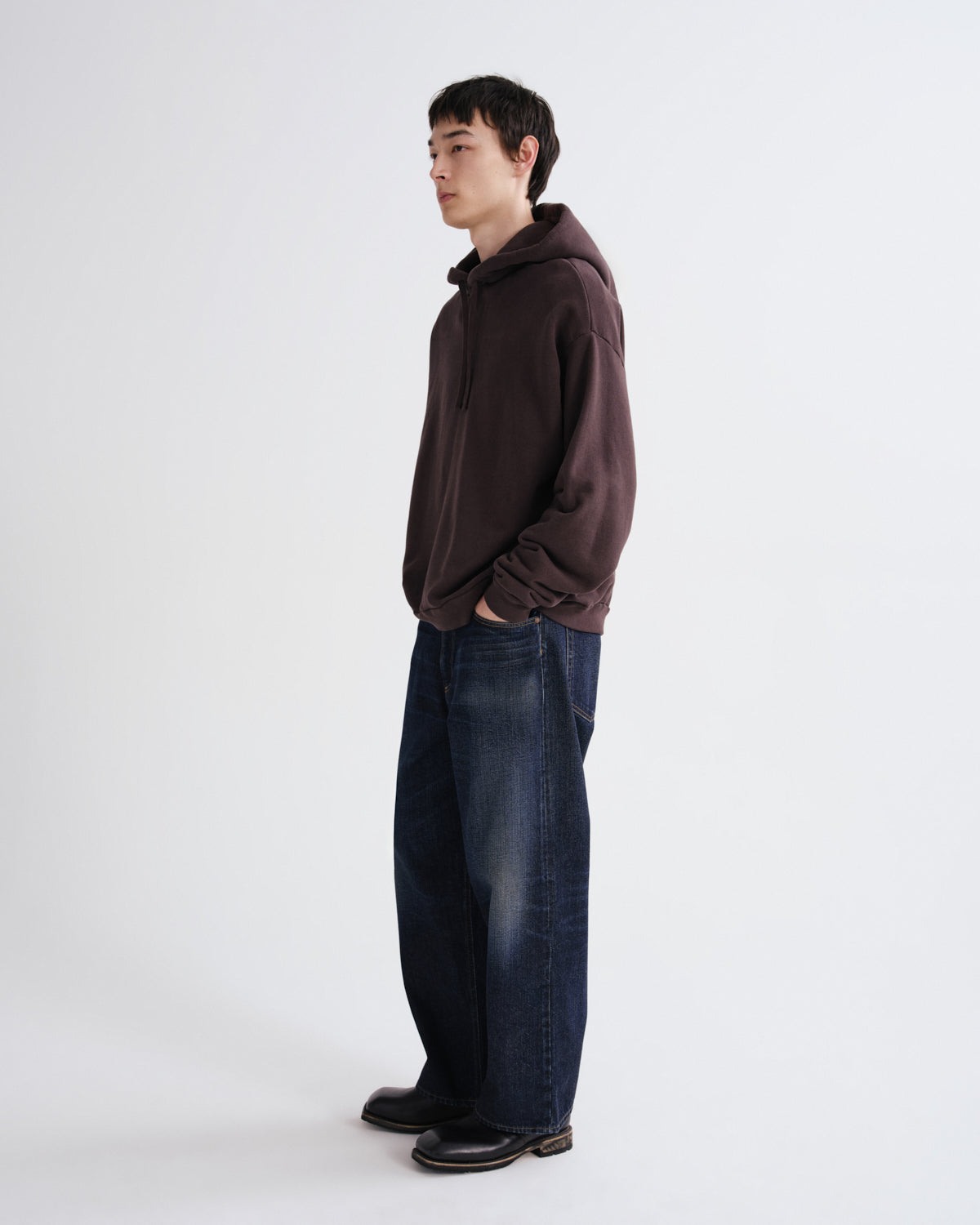 A man standing in profile wearing a dark brown oversized hoodie, loose dark blue jeans, and black shoes against a plain light gray studio background.