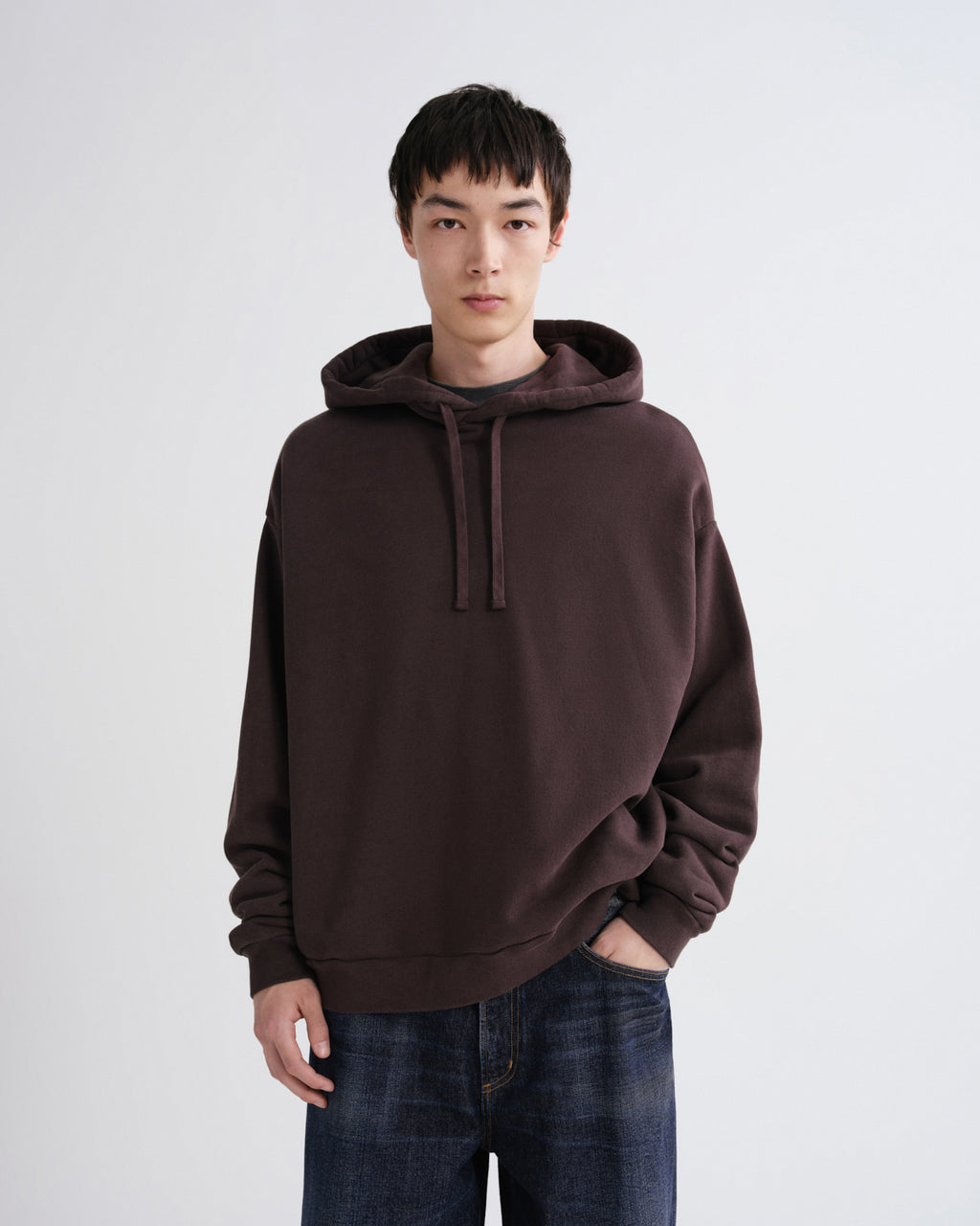 A man wearing an oversized dark brown pullover hoodie with drawstrings and blue jeans, standing against a light gray studio background.