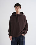 A man wearing an oversized dark brown pullover hoodie with drawstrings and blue jeans, standing against a light gray studio background.