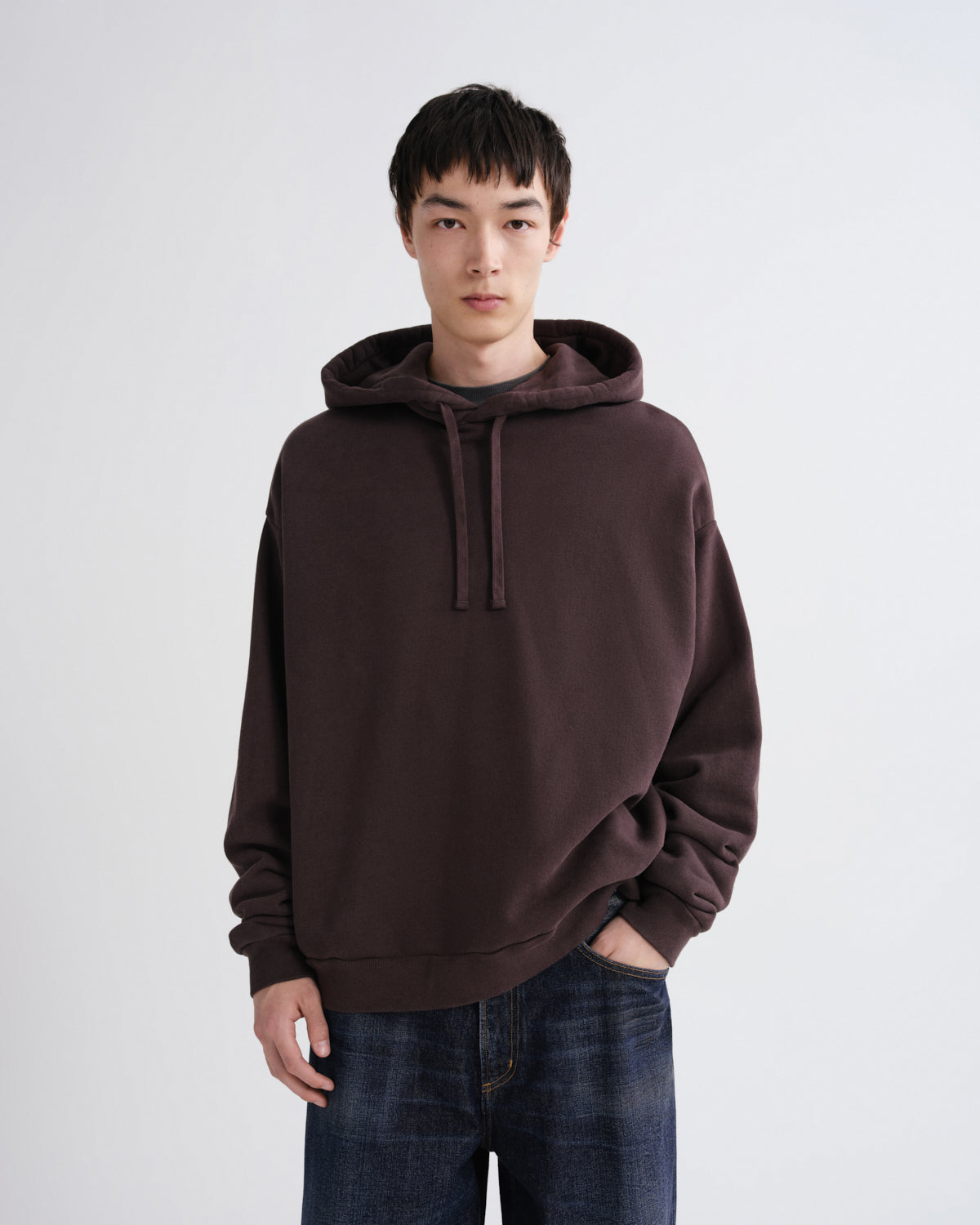 A man wearing an oversized dark brown pullover hoodie with drawstrings and blue jeans, standing against a light gray studio background.