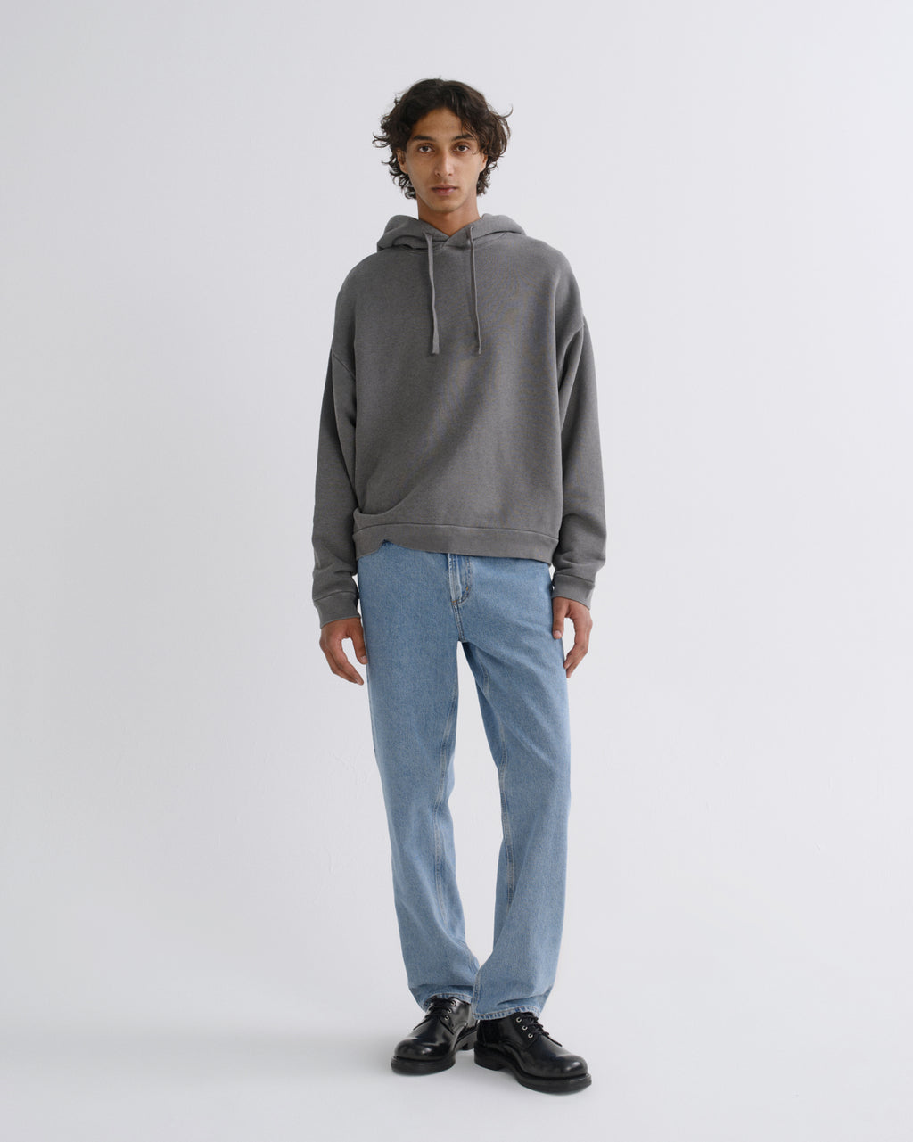 A model stands against a light gray studio background wearing a dark gray pullover hoodie, light blue jeans, and black leather shoes.