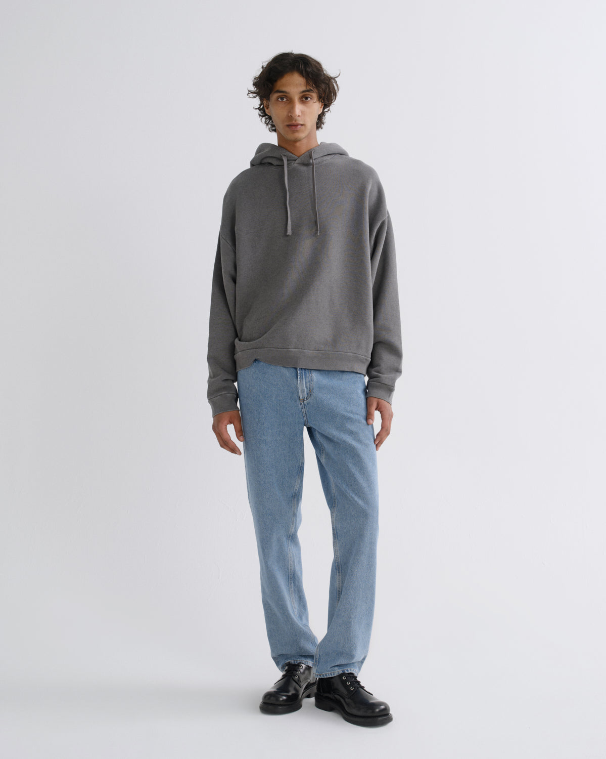 A model stands against a light gray studio background wearing a dark gray pullover hoodie, light blue jeans, and black leather shoes.