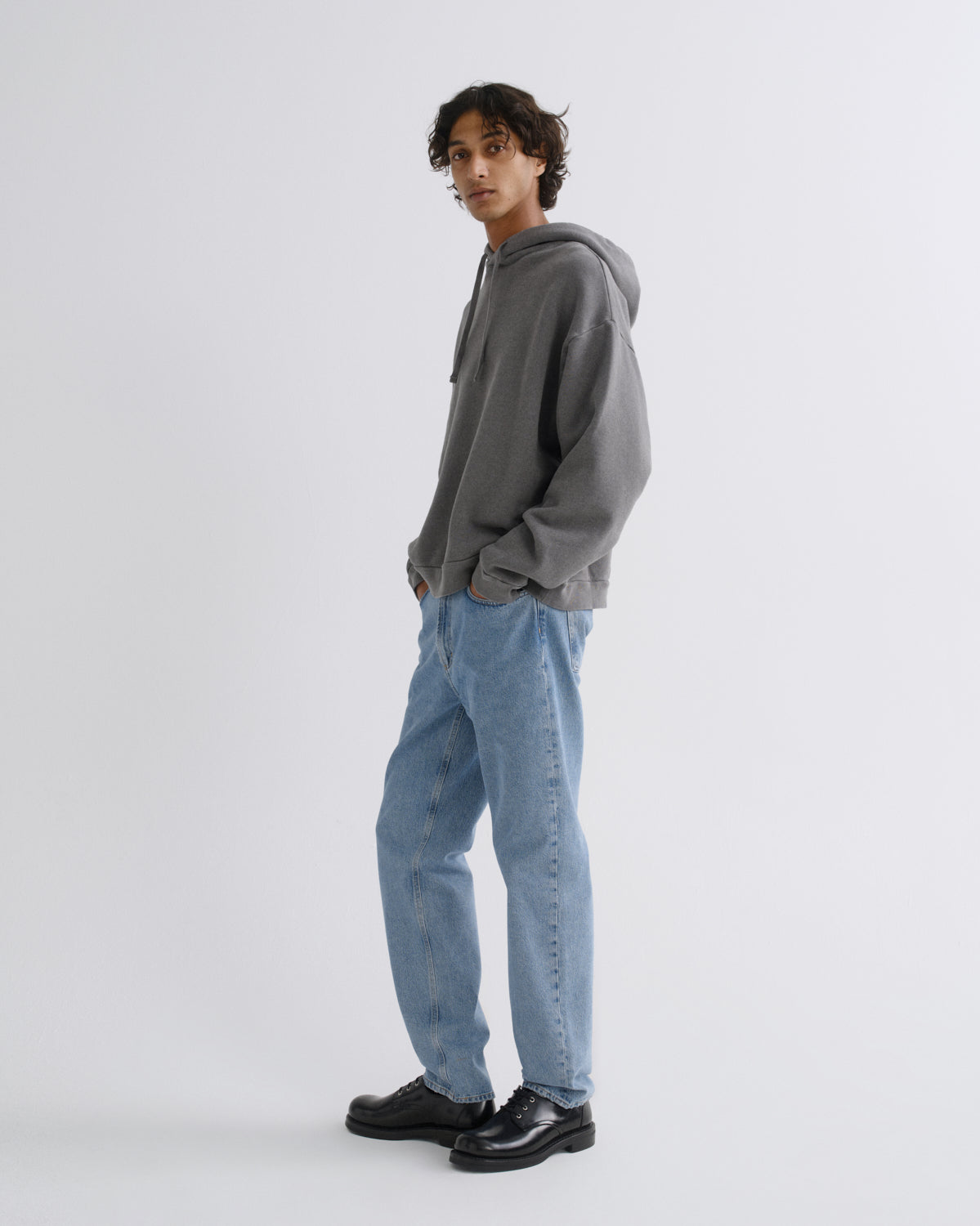 A man wearing a gray oversized hoodie, light blue jeans, and black leather shoes, standing in profile against a light gray studio background.