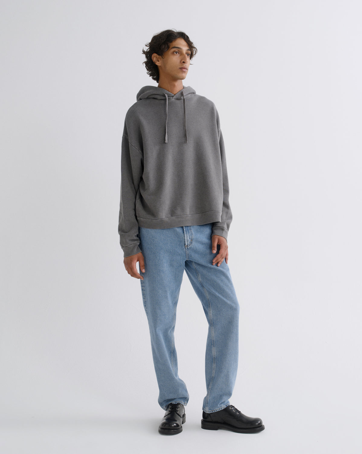 A man wearing a dark gray oversized hoodie, light blue jeans, and black leather shoes, standing against a light gray studio background.