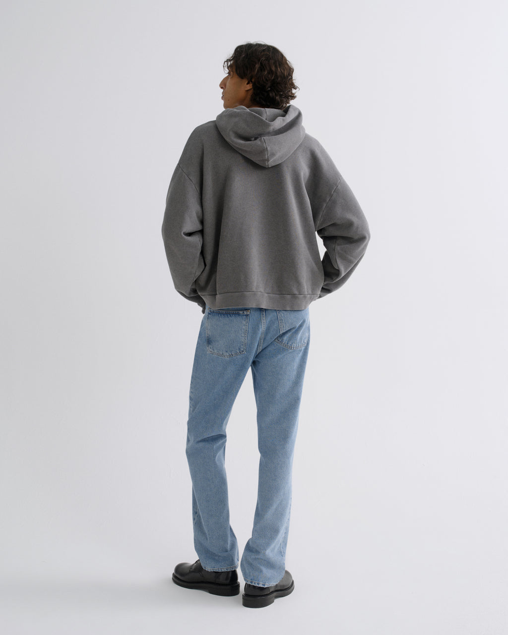 A model shown from the back wearing a washed gray oversized hoodie, light blue jeans, and black shoes, standing against a light gray studio background.