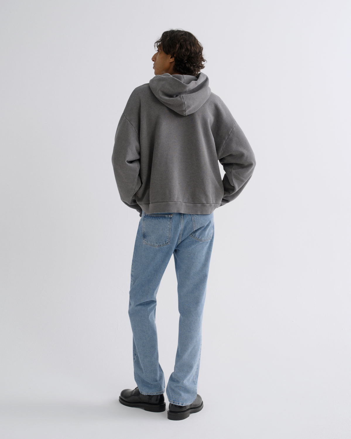 A model shown from the back wearing a washed gray oversized hoodie, light blue jeans, and black shoes, standing against a light gray studio background.