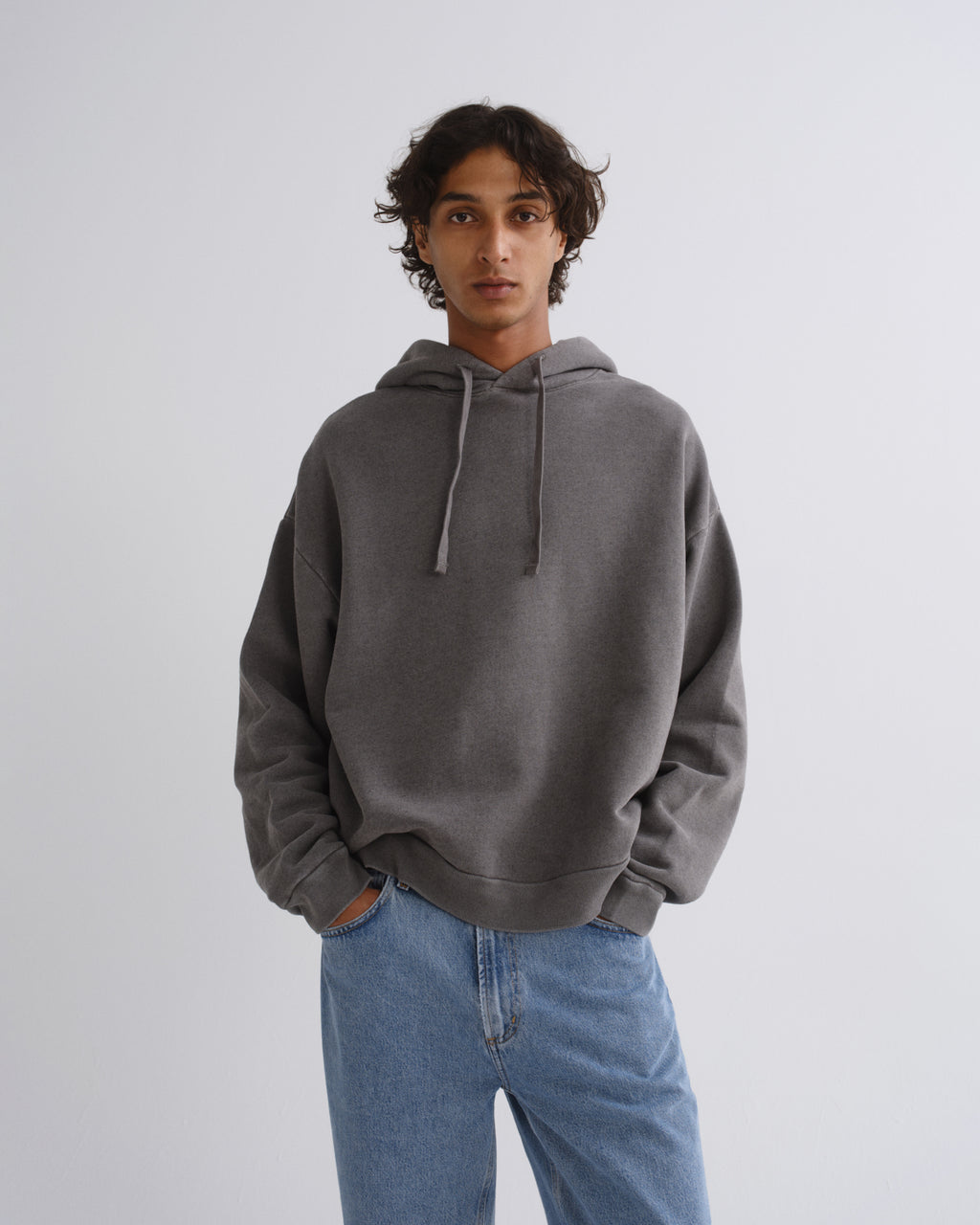 A man wearing a dark gray oversized pullover hoodie with matching drawstrings and light blue jeans, standing against a light gray studio background.