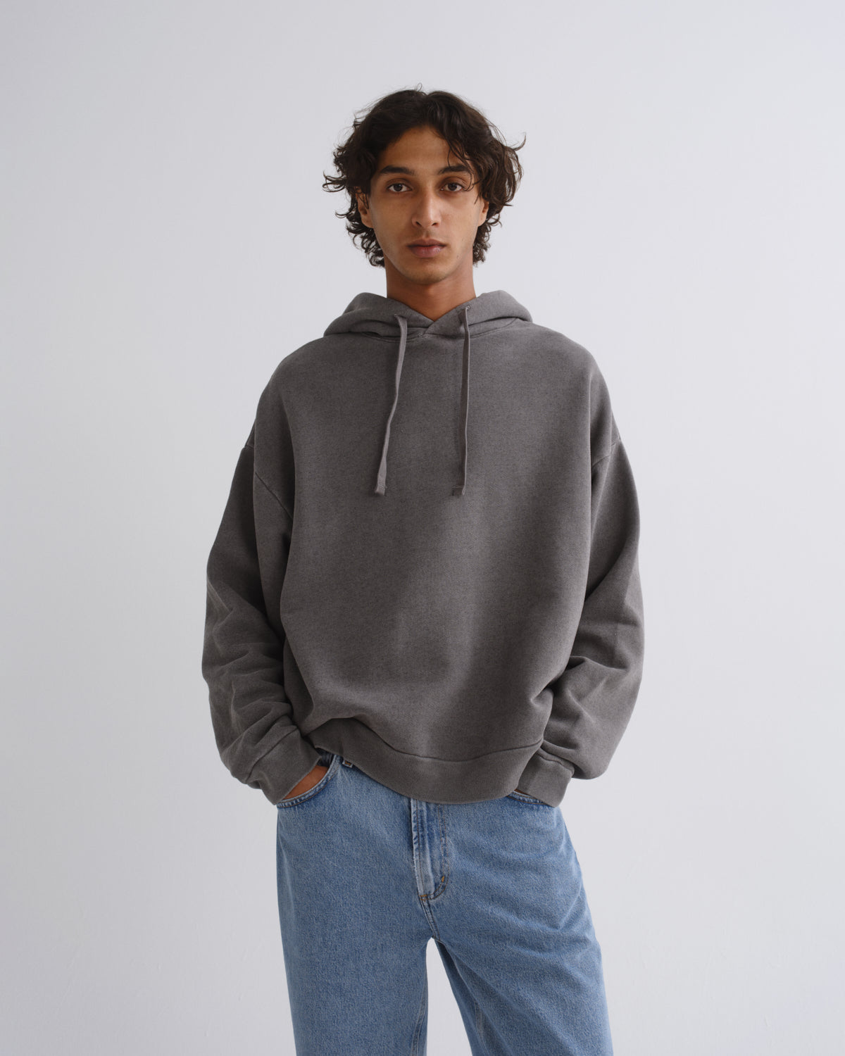 A man wearing a dark gray oversized pullover hoodie with matching drawstrings and light blue jeans, standing against a light gray studio background.