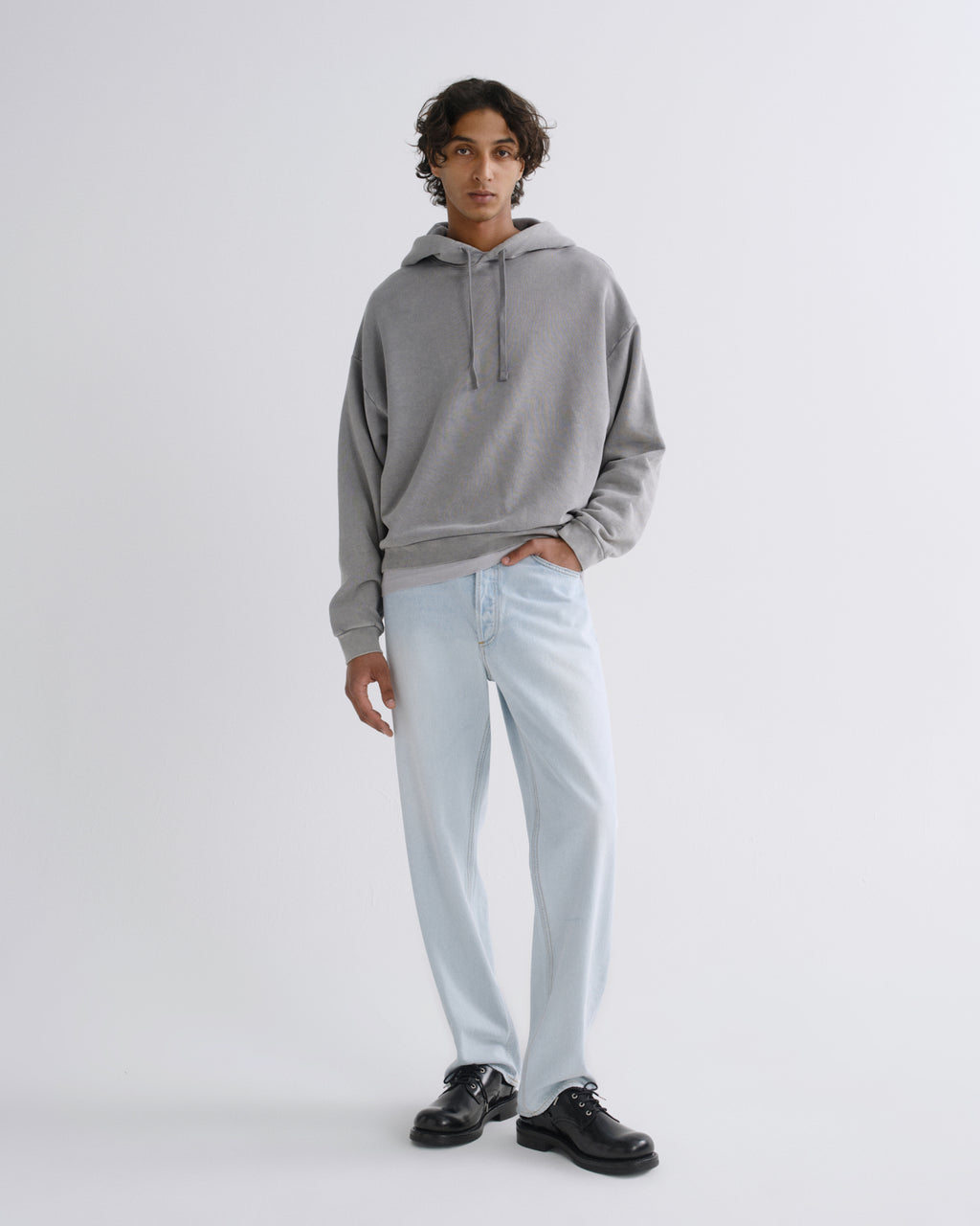 A man modeling a light gray oversized hoodie, light-wash blue jeans, and black leather shoes against a plain light gray studio background.