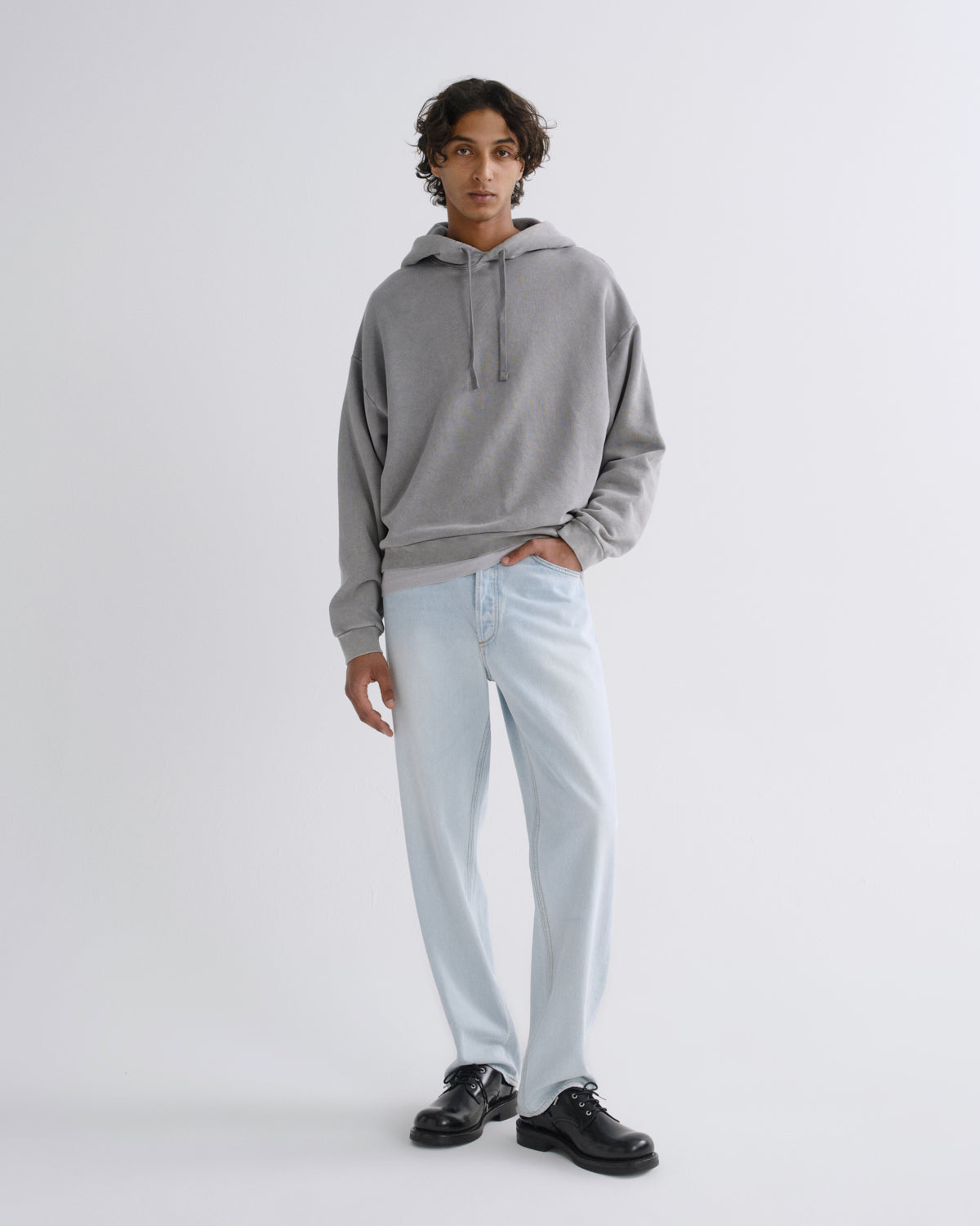 A man modeling a light gray oversized hoodie, light-wash blue jeans, and black leather shoes against a plain light gray studio background.