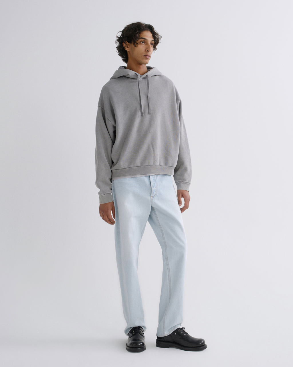 A man modeling a light gray pullover hoodie with light-wash blue jeans and black leather shoes, standing against a plain light gray studio background.
