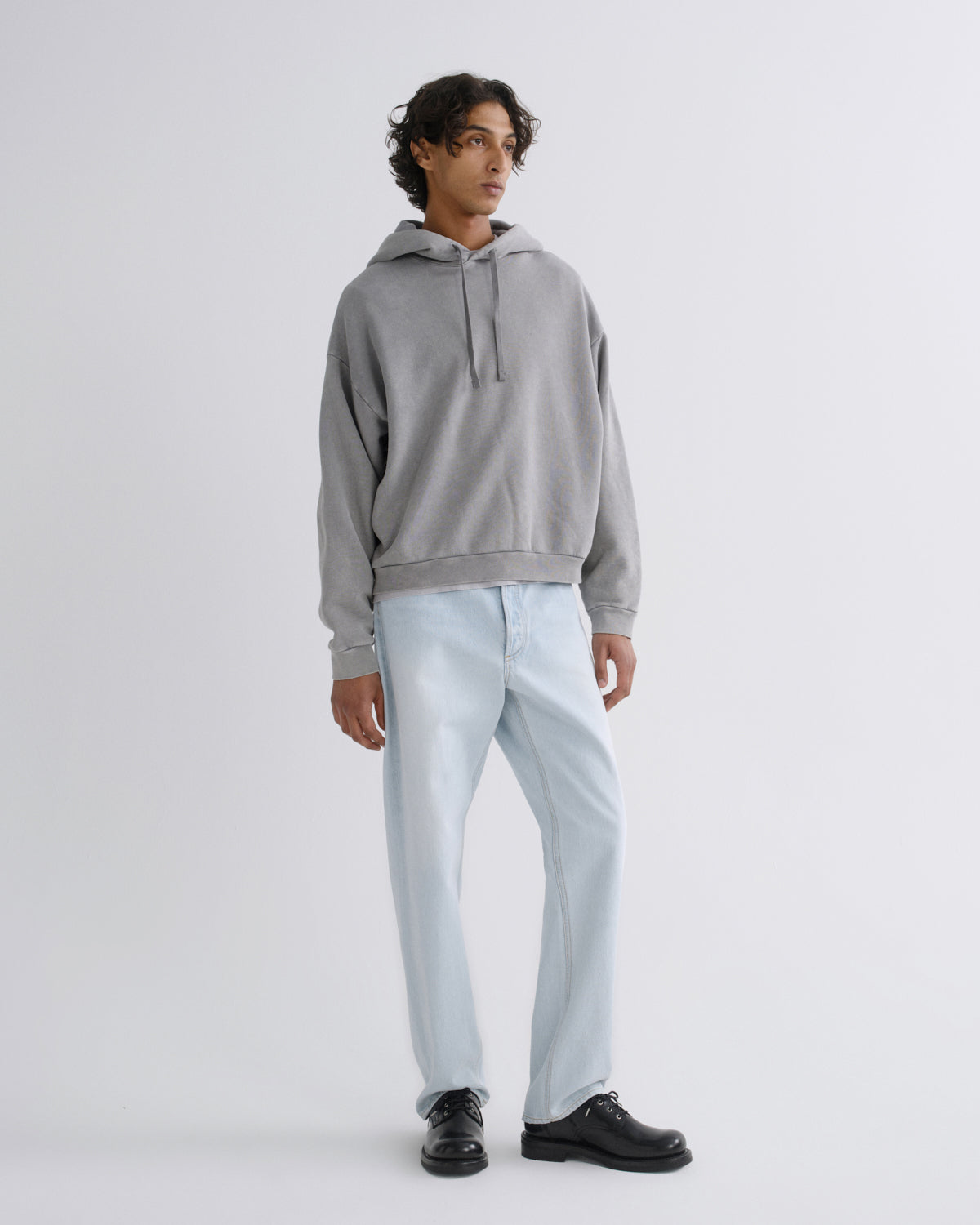 A man modeling a light gray pullover hoodie with light-wash blue jeans and black leather shoes, standing against a plain light gray studio background.