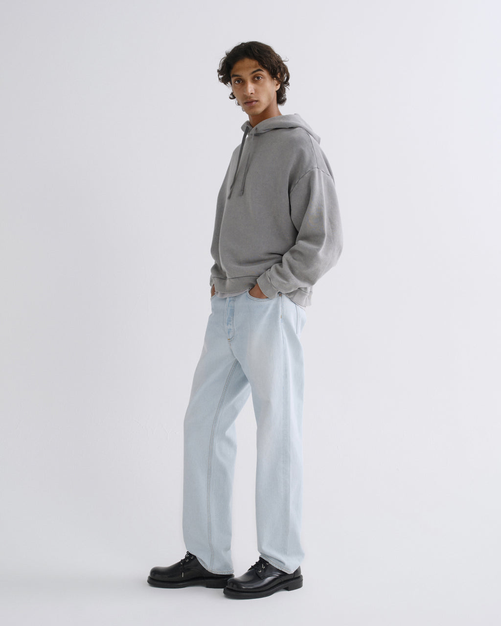 A model wearing a light gray oversized hoodie, light-wash wide-leg jeans, and black leather shoes, standing in profile against a plain light gray studio background.