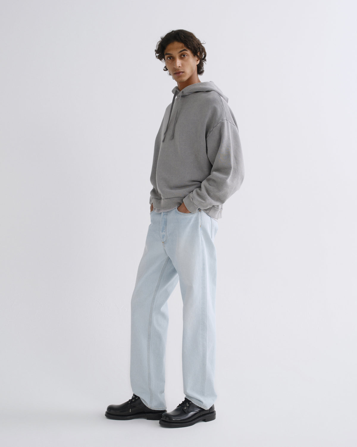 A model wearing a light gray oversized hoodie, light-wash wide-leg jeans, and black leather shoes, standing in profile against a plain light gray studio background.