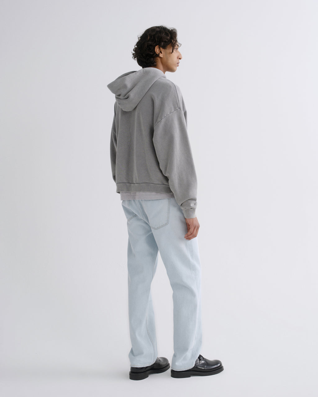 A model shown from the back wearing a gray washed hoodie, light wash jeans, and black leather shoes against a plain light gray studio background.