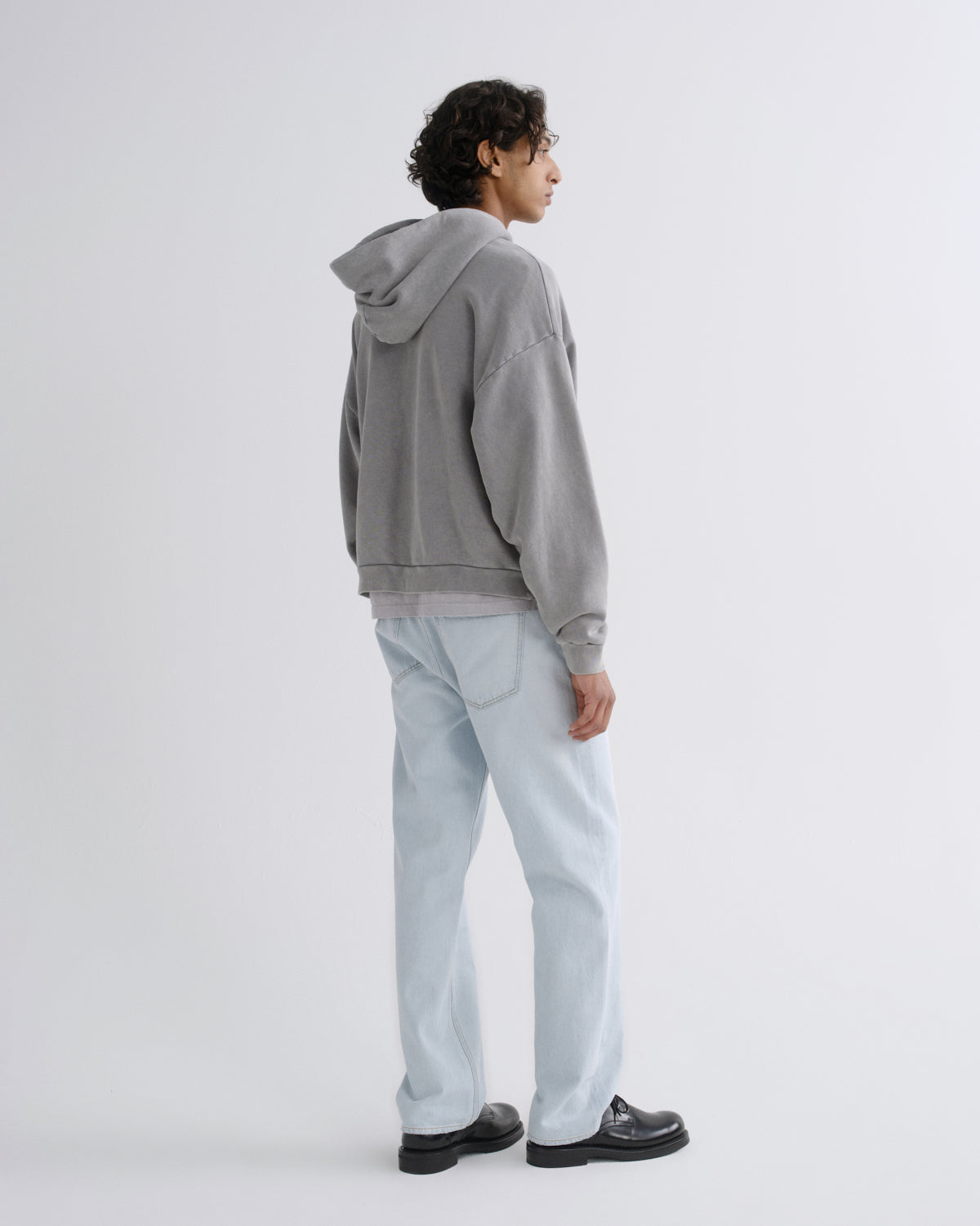 A model shown from the back wearing a gray washed hoodie, light wash jeans, and black leather shoes against a plain light gray studio background.