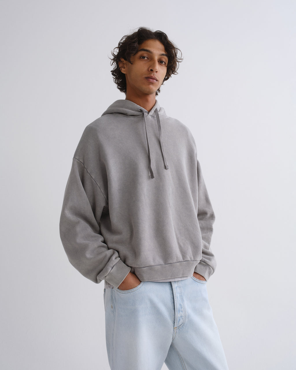A man wearing a light gray oversized pullover hoodie with drawstrings and light-wash blue jeans, standing against a plain light gray studio background.