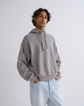 A man wearing a light gray oversized pullover hoodie with drawstrings and light-wash blue jeans, standing against a plain light gray studio background.