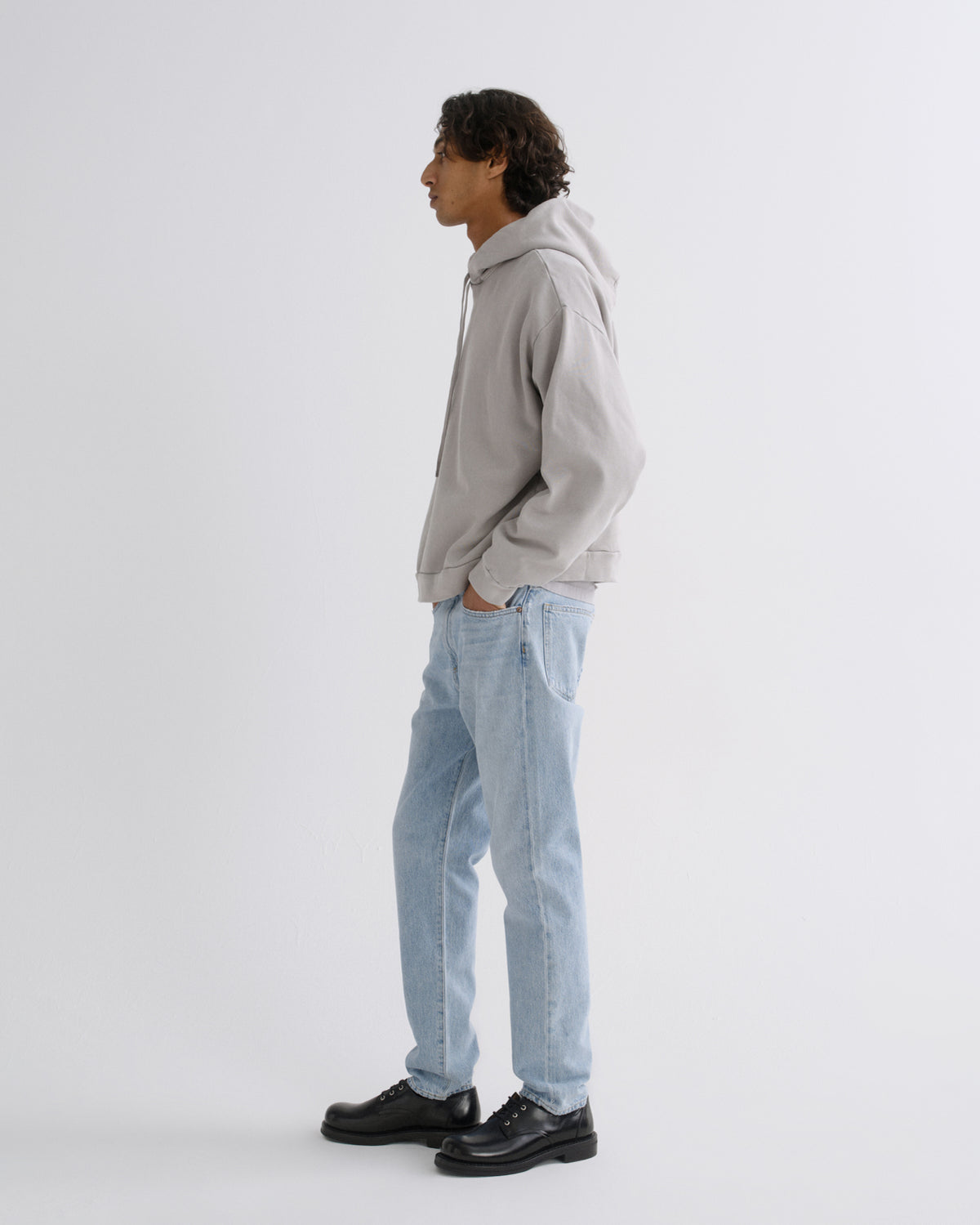 A man in a light gray hoodie, light blue jeans, and black leather shoes stands in profile against a plain light gray studio background.