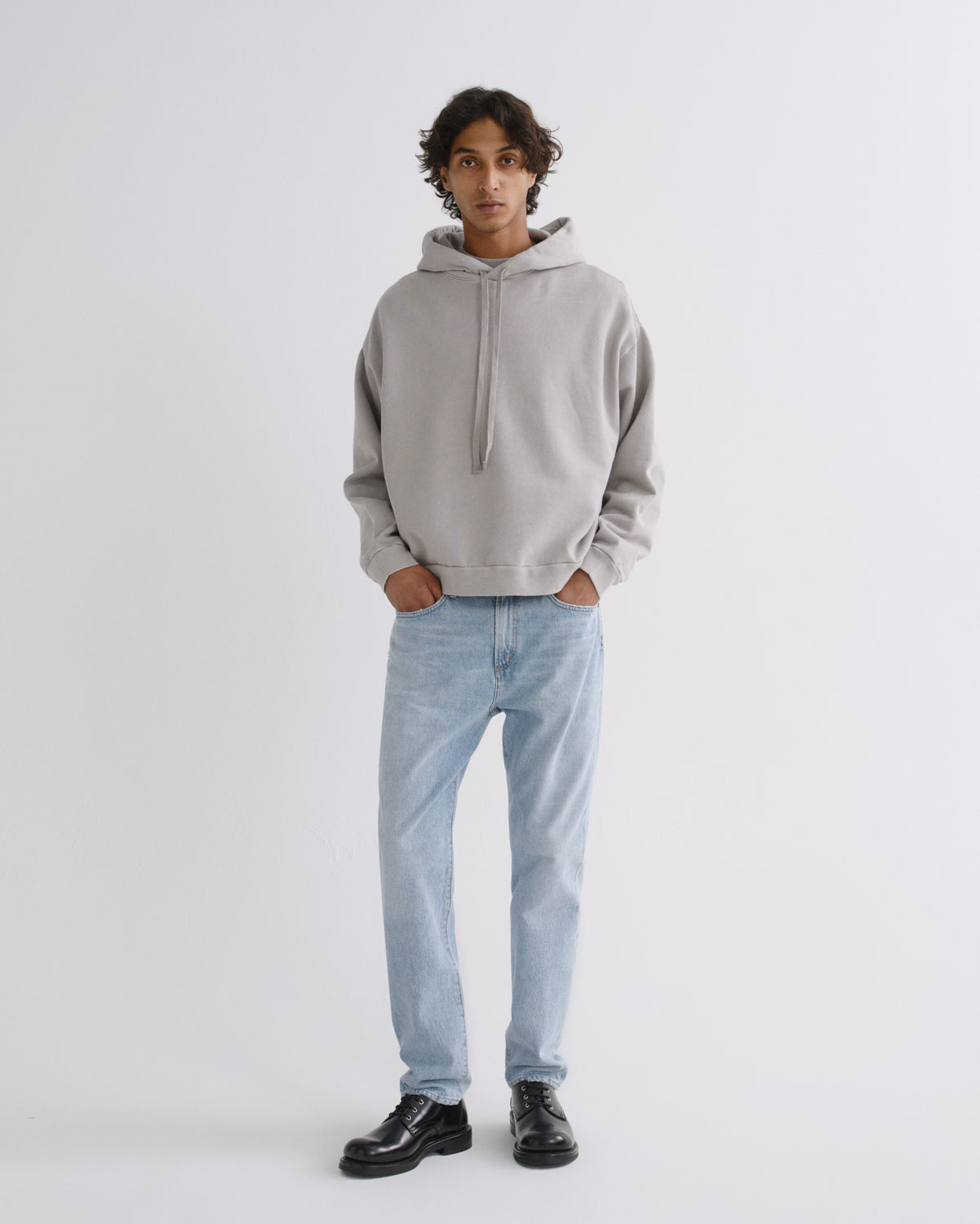 A model stands against a light gray studio background wearing a light gray oversized hoodie, light blue jeans, and black leather shoes.