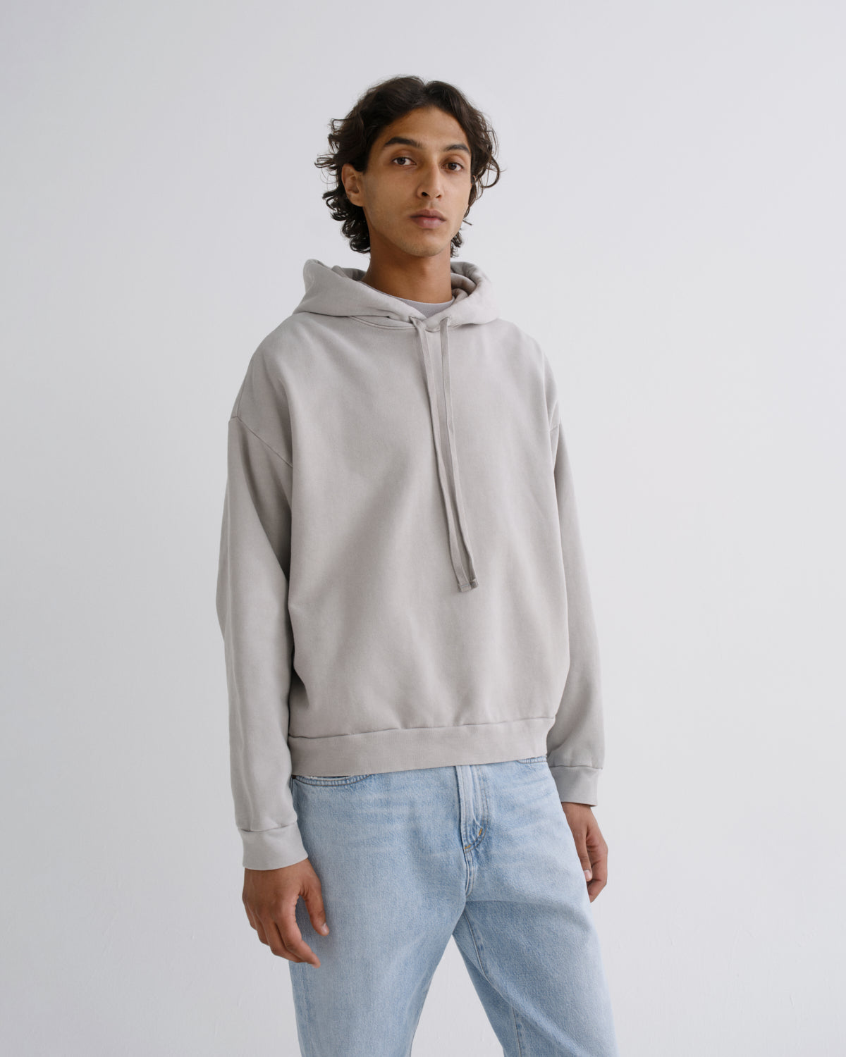 A man wearing a light gray oversized pullover hoodie with matching drawstrings and light blue jeans, standing against a plain light gray studio background.