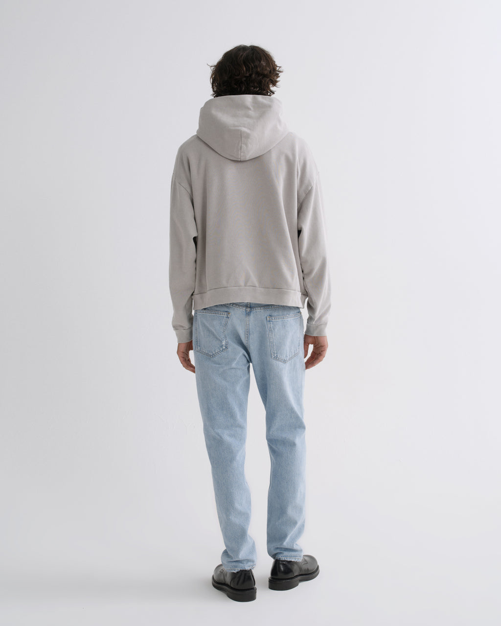 A person shown from the back wearing a light gray oversized hoodie and light blue jeans, standing against a plain light gray studio background.