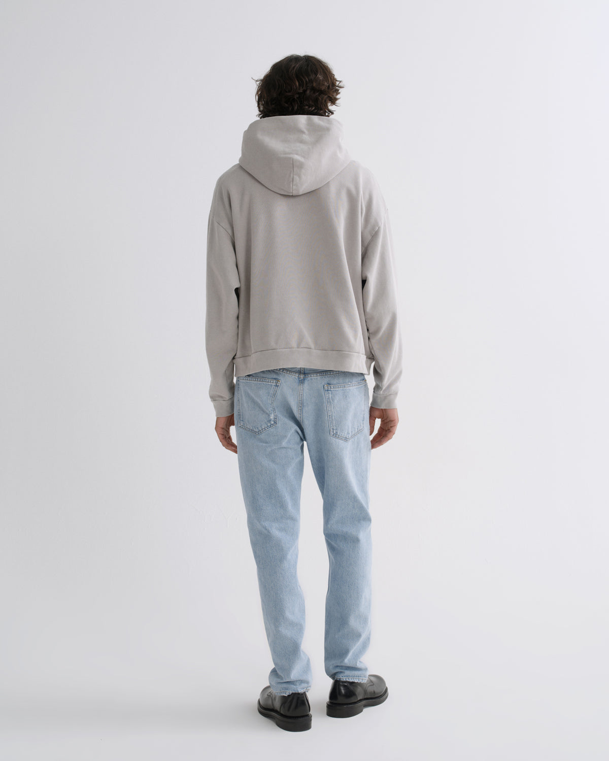 A person shown from the back wearing a light gray oversized hoodie and light blue jeans, standing against a plain light gray studio background.