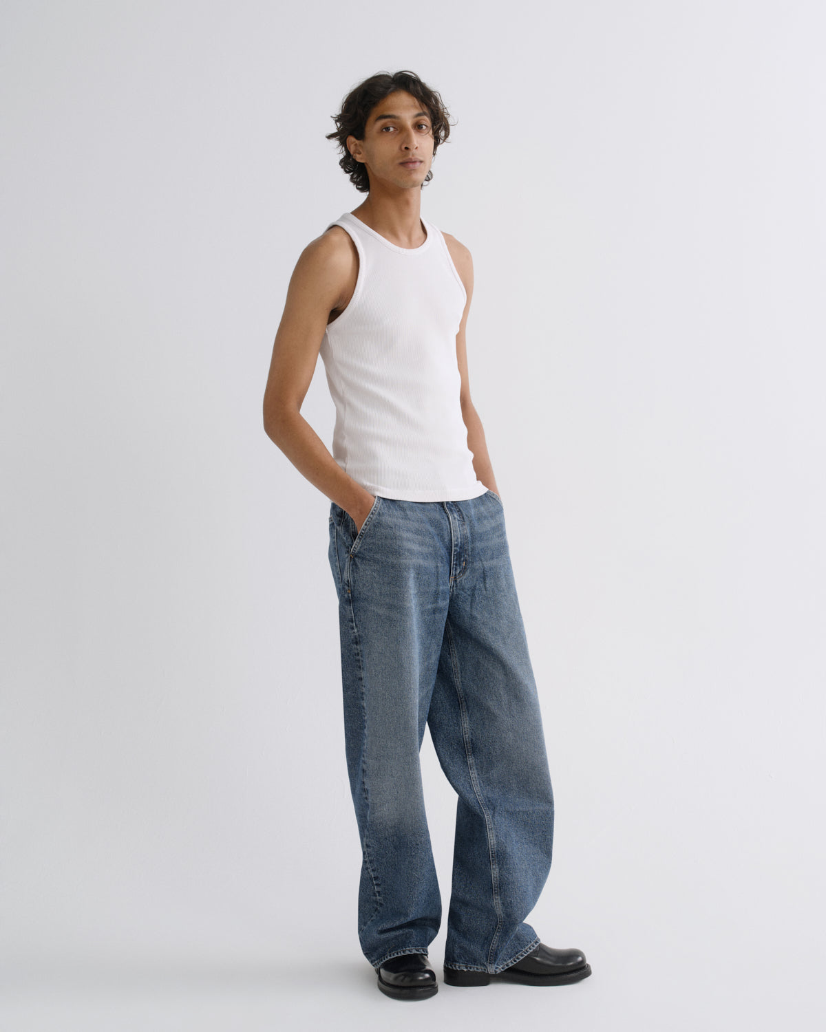 A man modeling a white ribbed tank top and wide-leg blue jeans against a light gray studio background.