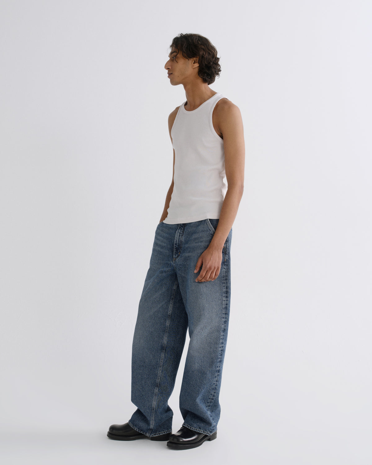 A model standing in profile wearing a white ribbed tank top, wide-leg blue jeans, and black leather shoes against a plain light gray studio background.