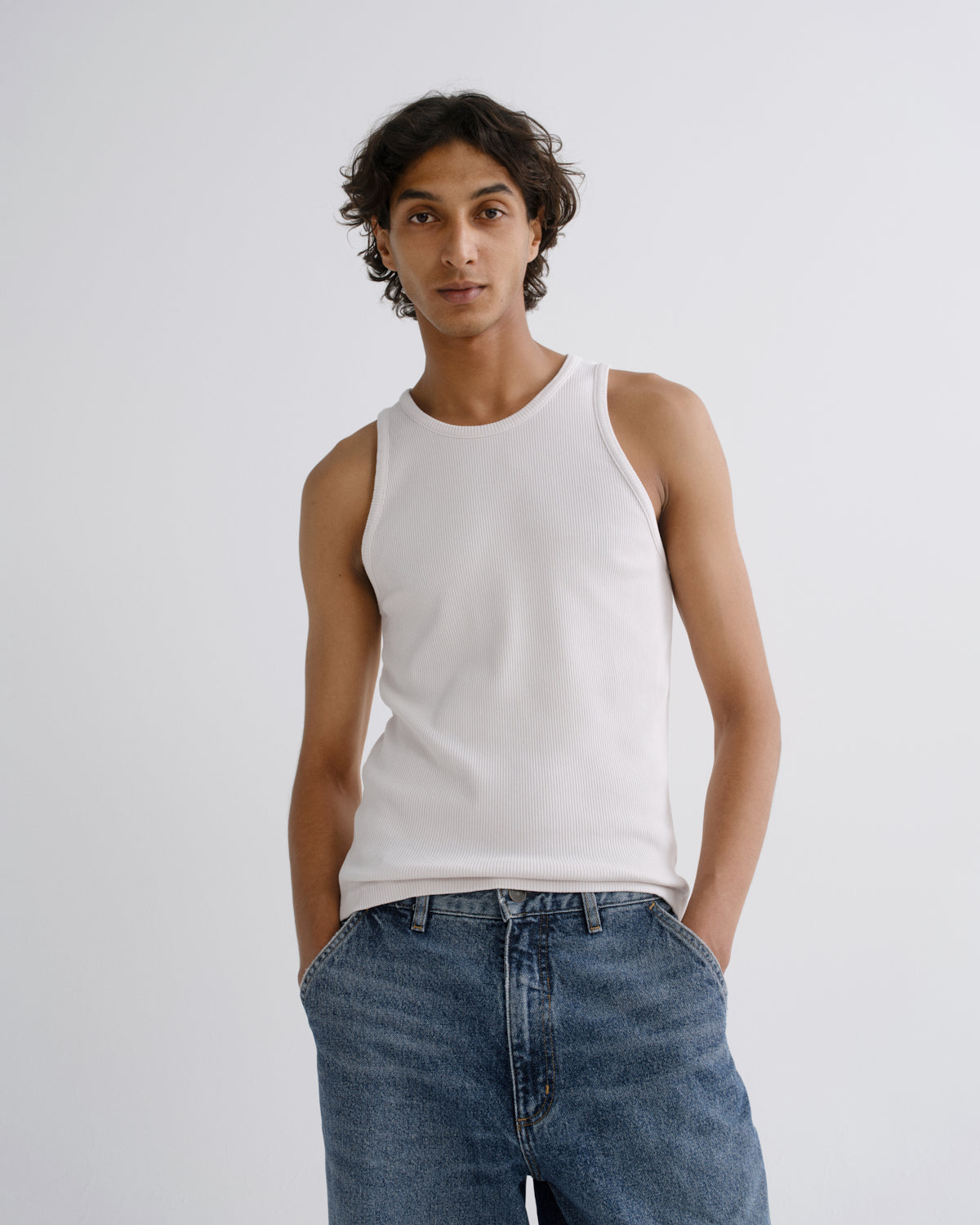 A man wearing a white ribbed tank top and blue jeans, standing against a light gray studio background.