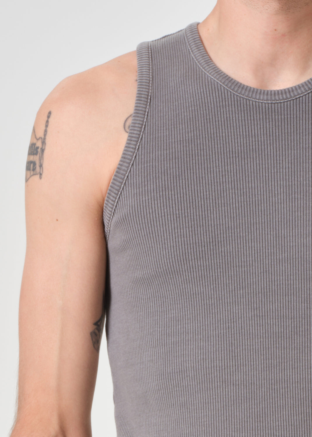 Person wearing a grey ribbed tank top with tattoos on arms against a grey background