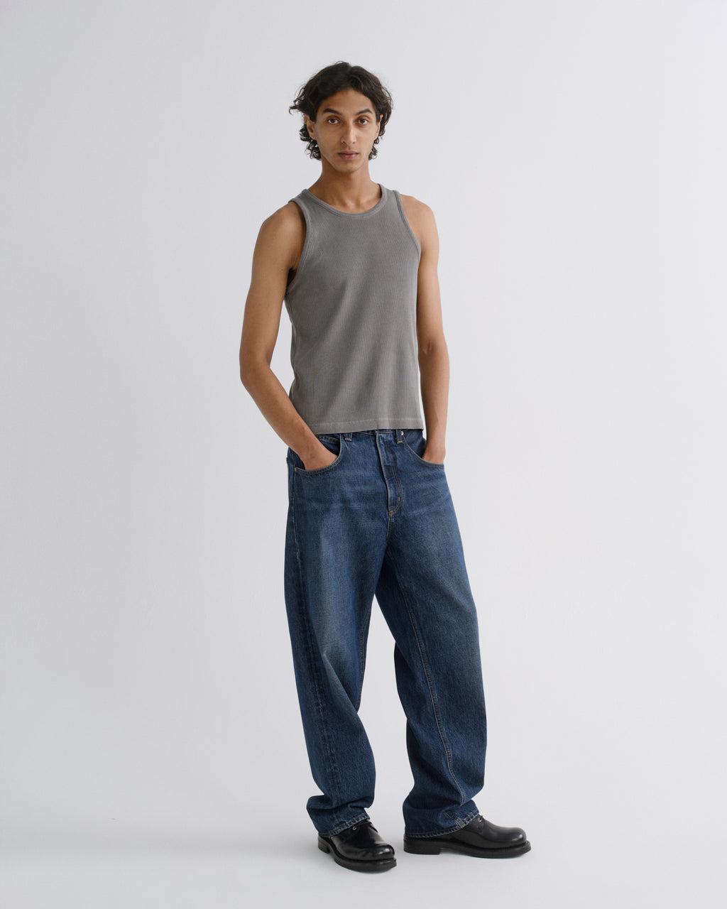 A model stands against a light gray studio background wearing a fitted gray sleeveless tank top, loose dark blue jeans, and black shoes.