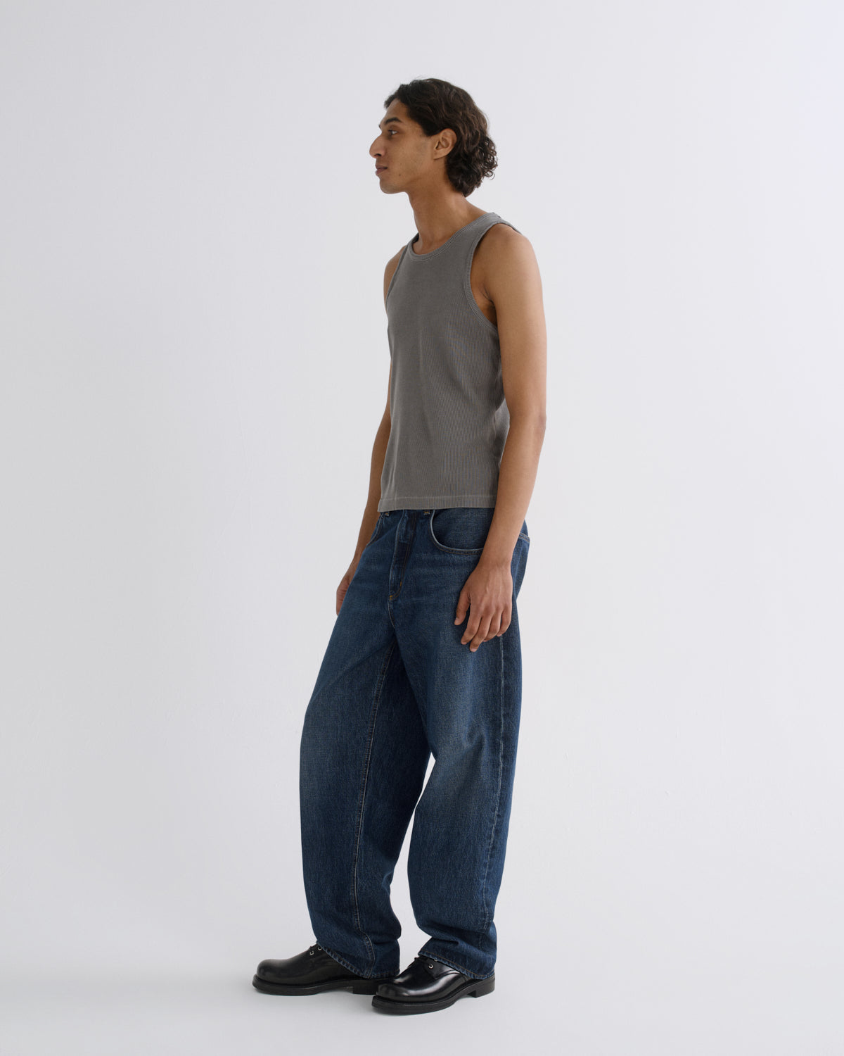 A man stands in profile wearing a gray sleeveless knit top, loose dark blue jeans, and black shoes against a plain light gray studio background.