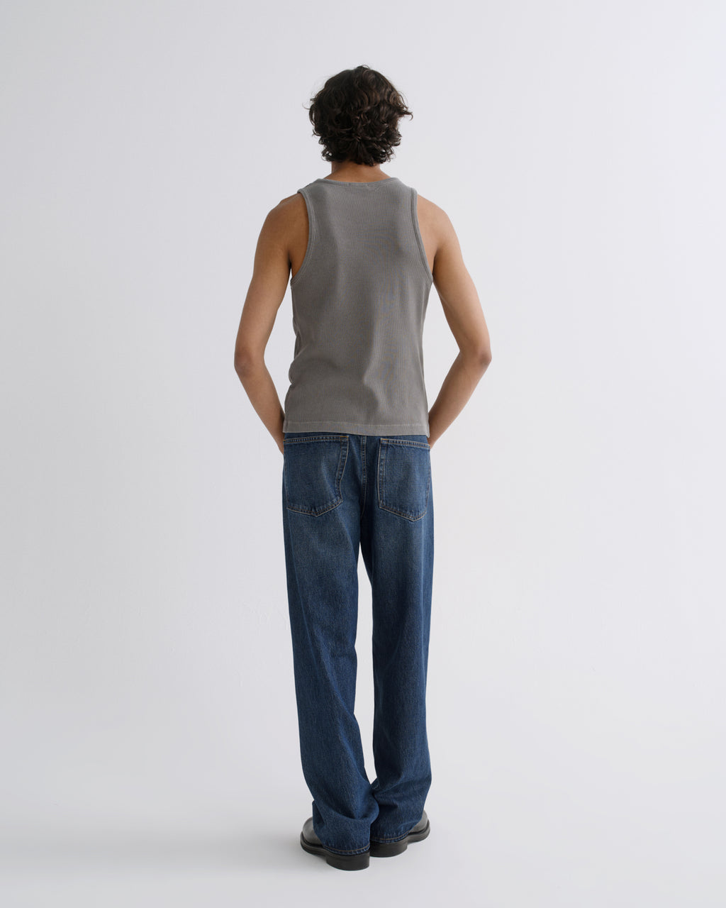 A man viewed from behind wearing a gray ribbed tank top and blue jeans against a light gray studio background.