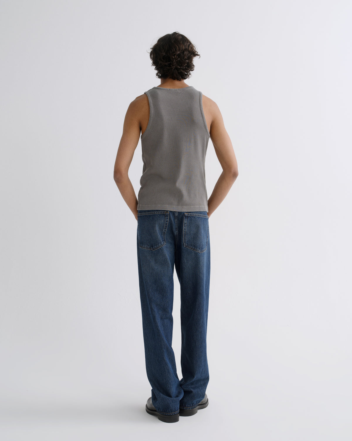 A man viewed from behind wearing a gray ribbed tank top and blue jeans against a light gray studio background.