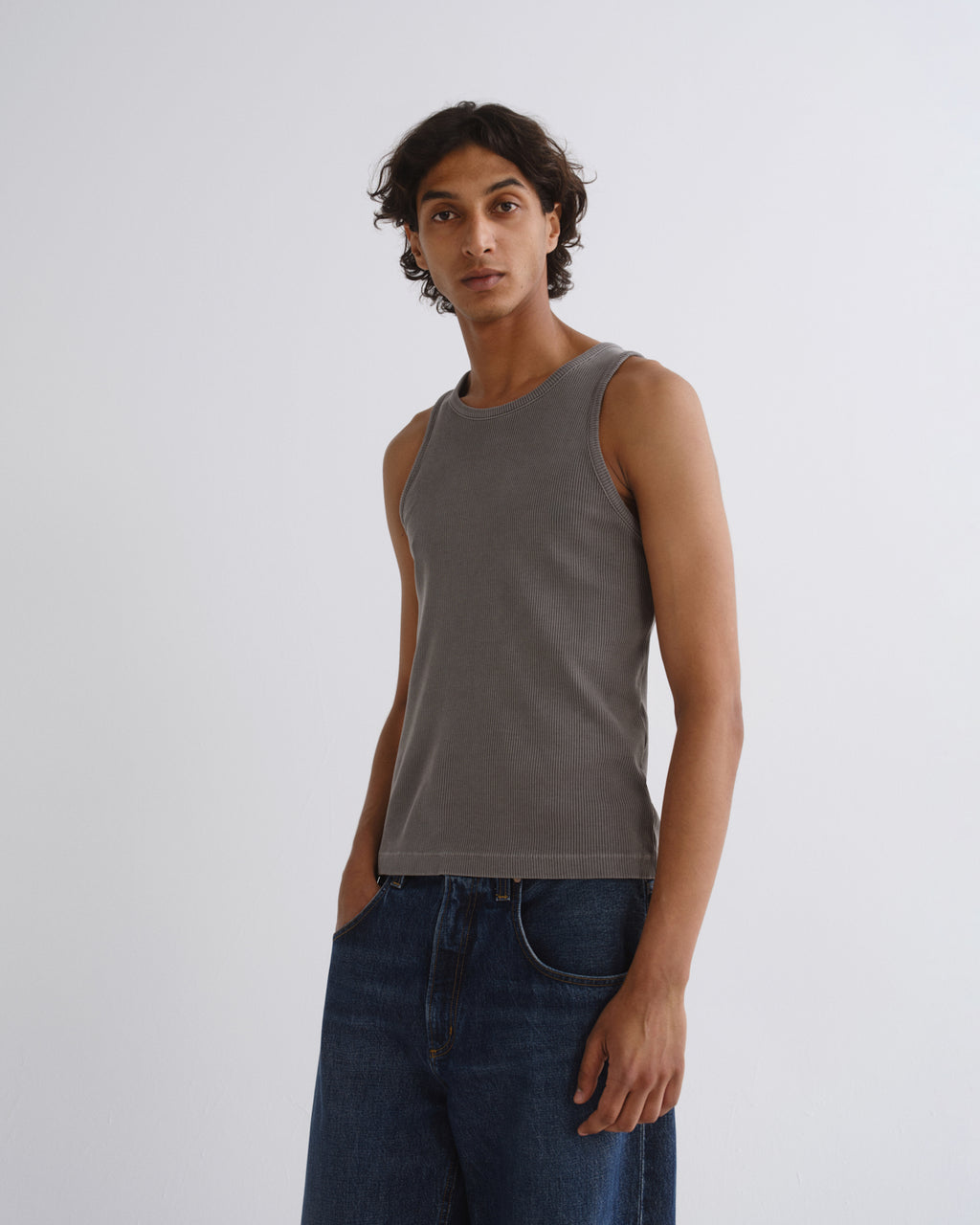 A man wearing a dark gray ribbed tank top and dark blue jeans, standing against a light gray studio background.