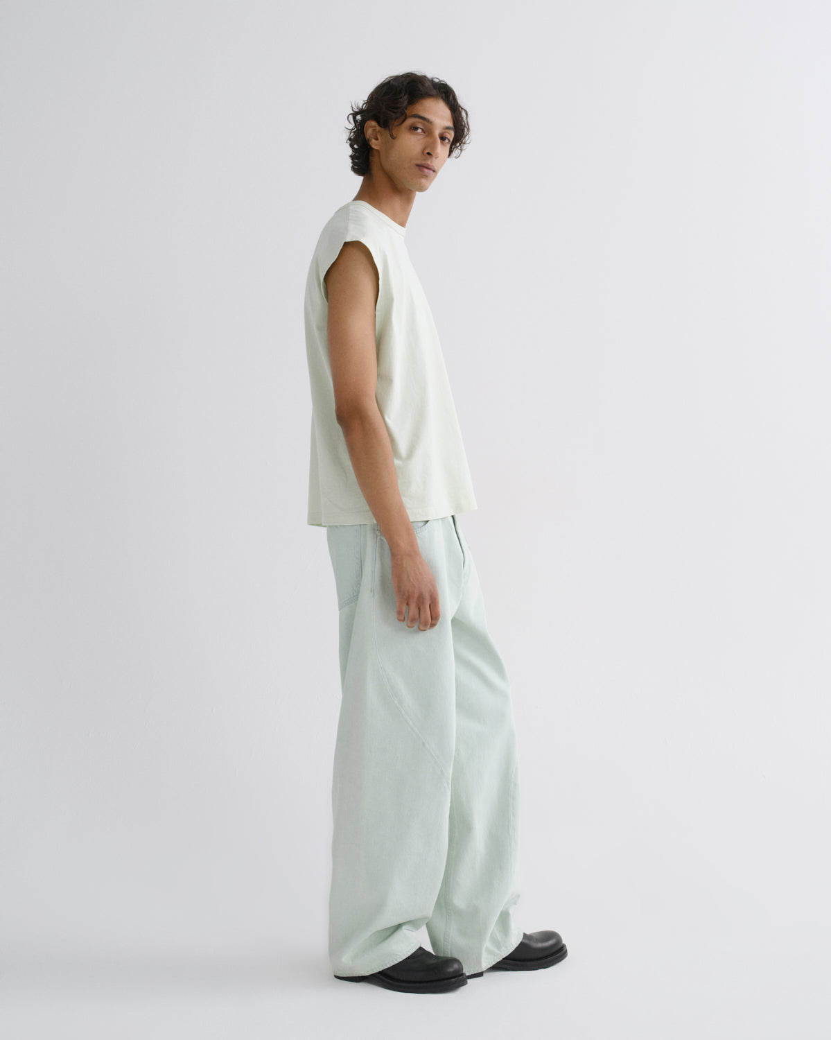 A man wearing a sleeveless pale green top and matching wide-leg light green pants, standing in profile against a plain light gray studio background.